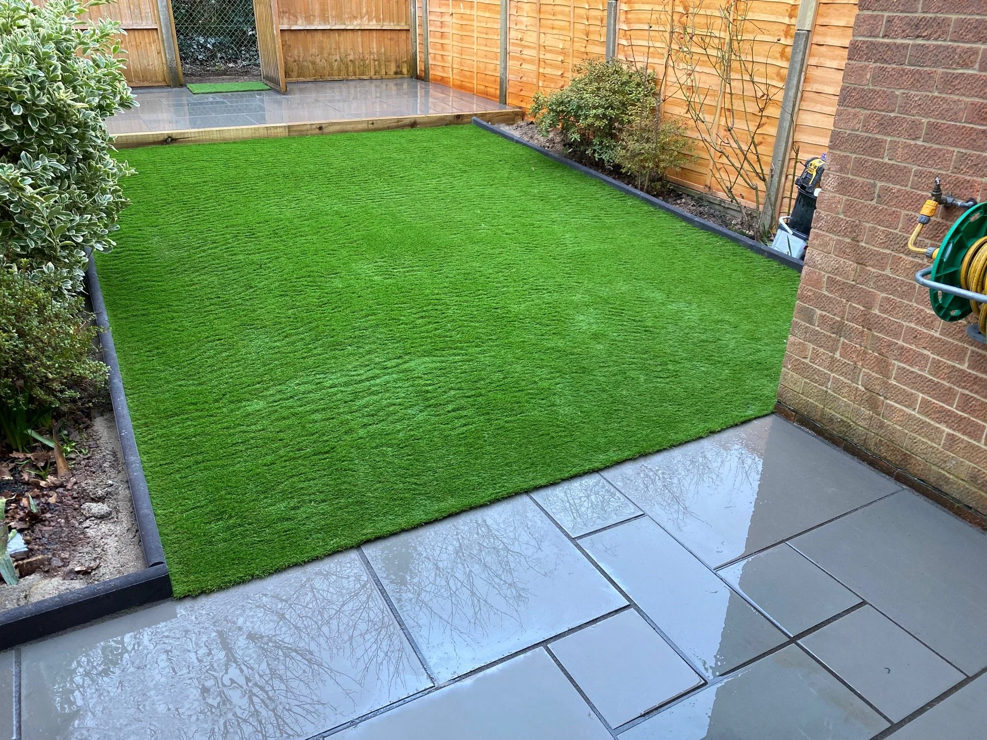 Artificial grass garden patch next to tiled patio with garden hose reel on brick wall