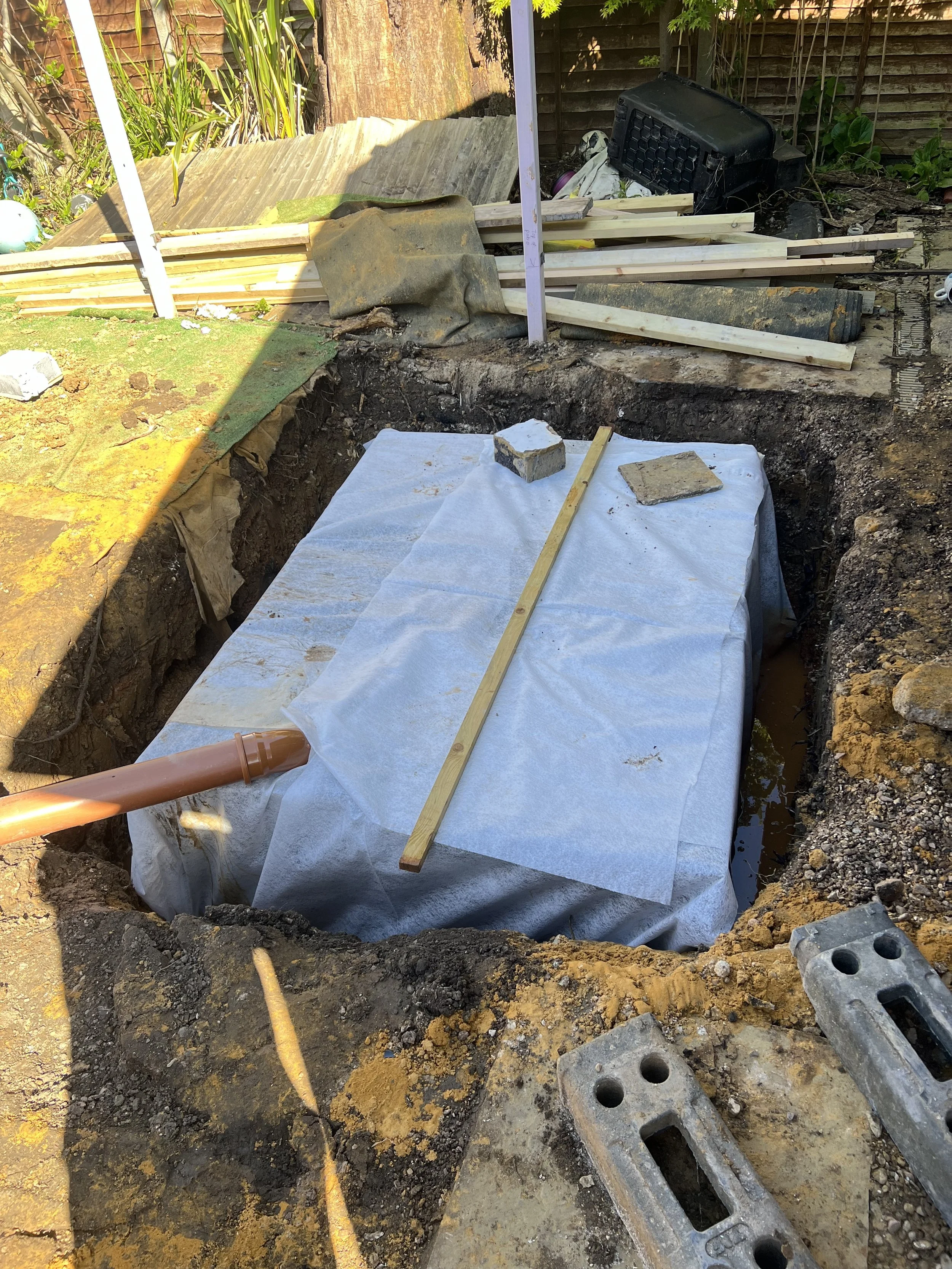 Construction site with a sunken area covered with a white tarp, pipes, wooden planks, bricks, and construction tools and materials.