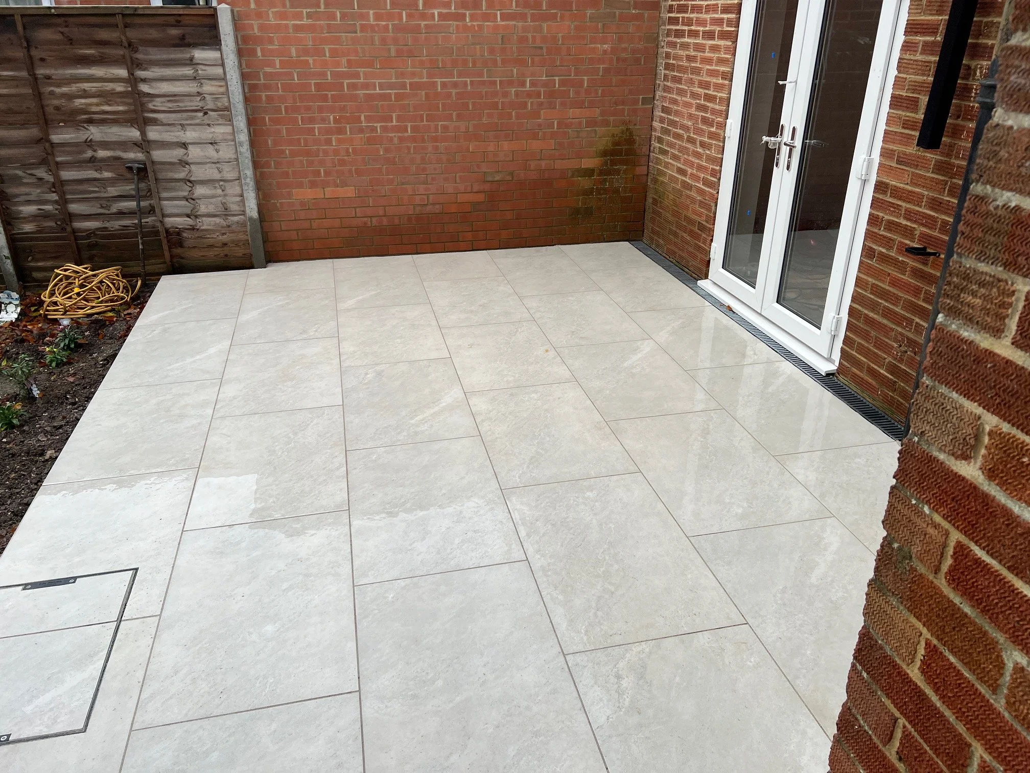 A newly paved outdoor patio area with large beige tiles, bordered by brick walls and a wooden fence, featuring a garden hose on the left and patio doors on the right.