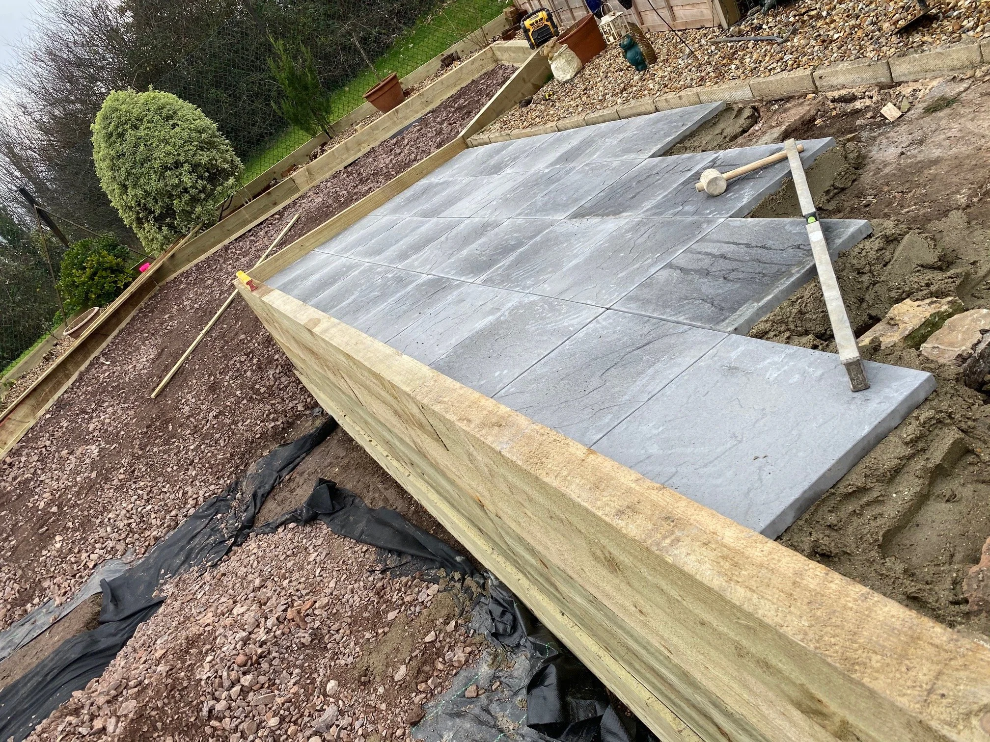 Construction site with new gray stone pavers laid on a wooden frame, on uneven ground with dirt and gravel, and tools resting on top.