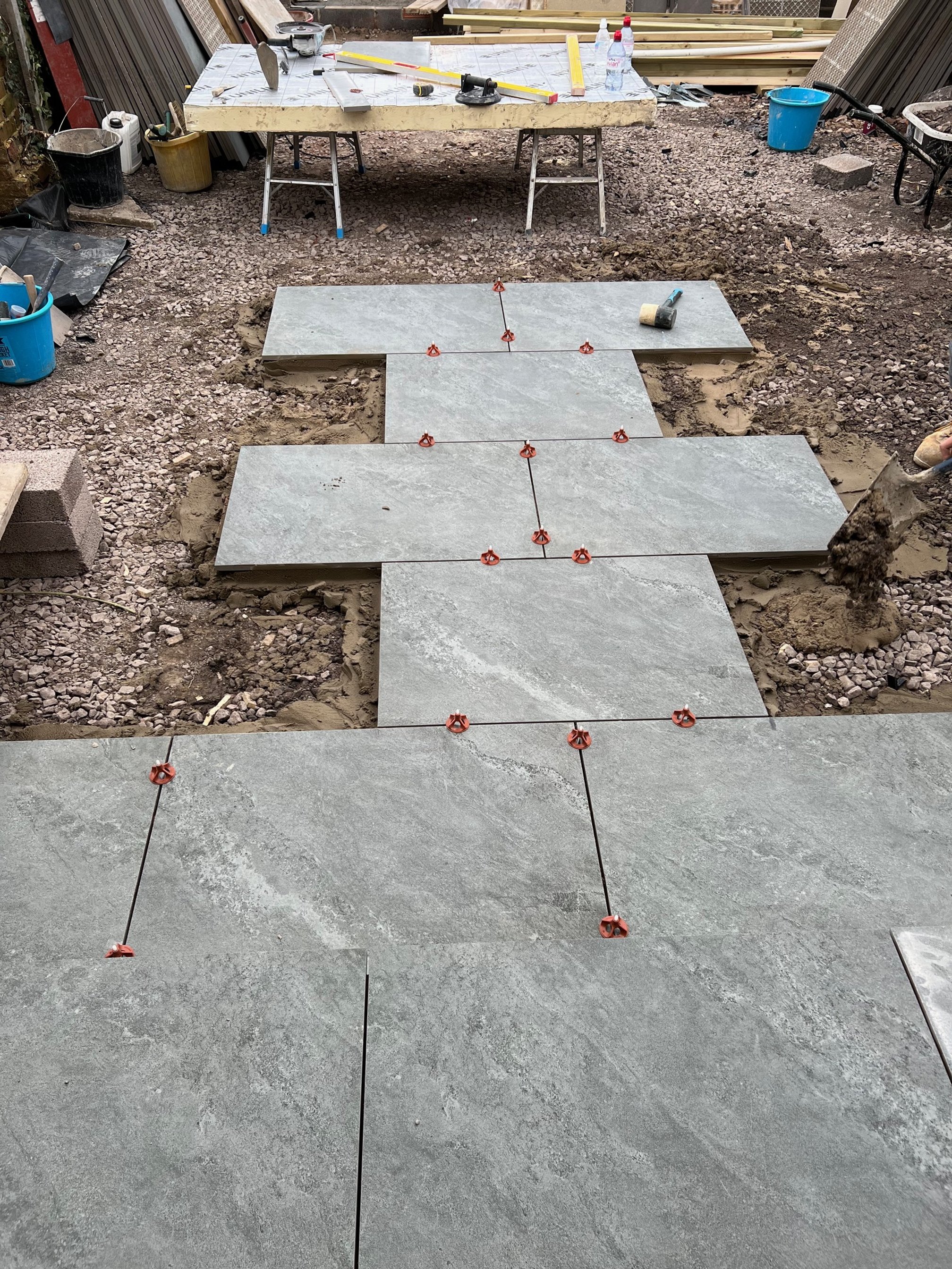 Laying large gray tiles on a construction site with tile spacers and a level on top, surrounded by gravel and construction tools.
