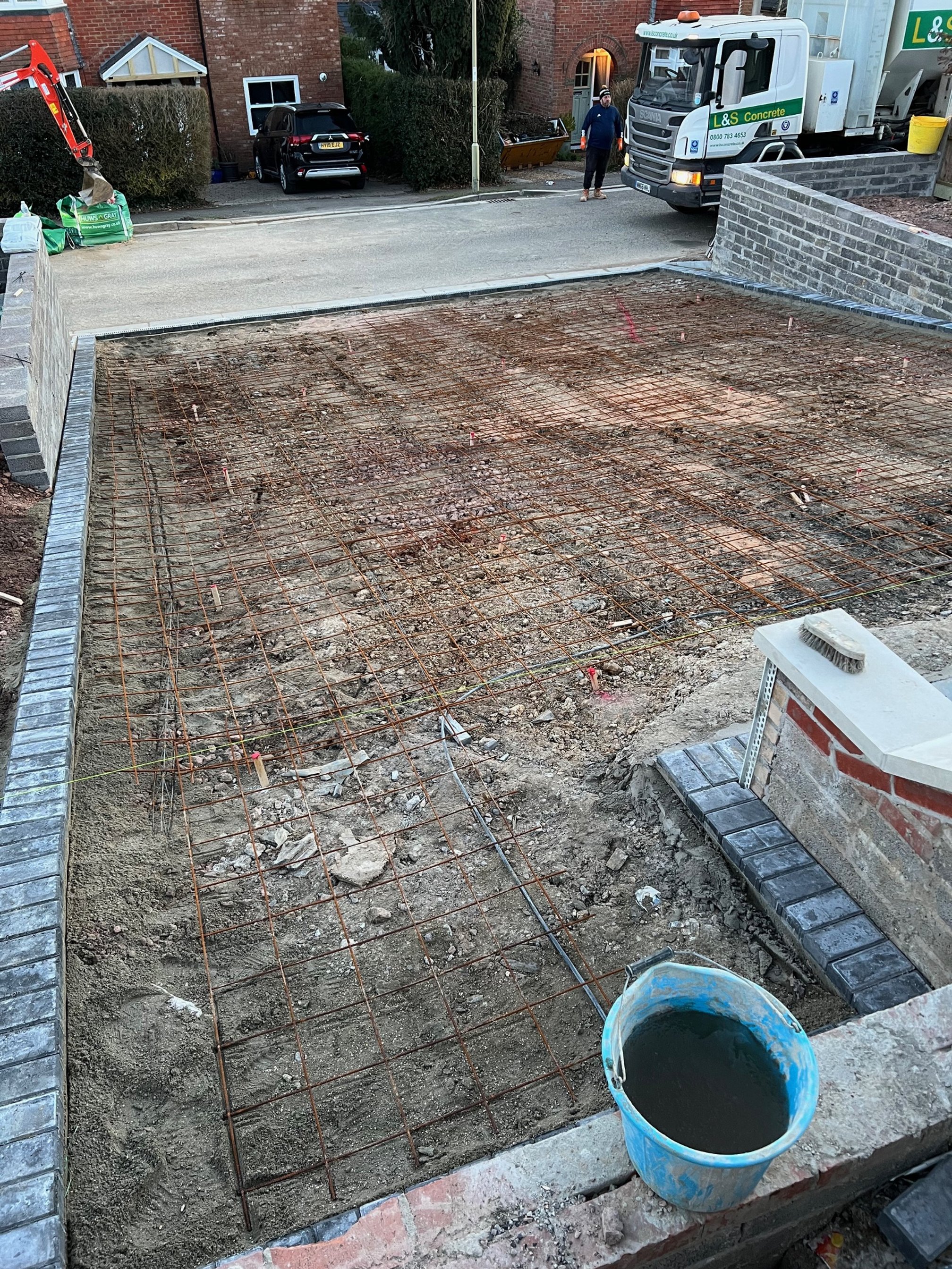 Construction site for a concrete driveway with rebar reinforcement, surrounded by brick and stone walls, and construction tools and materials nearby.