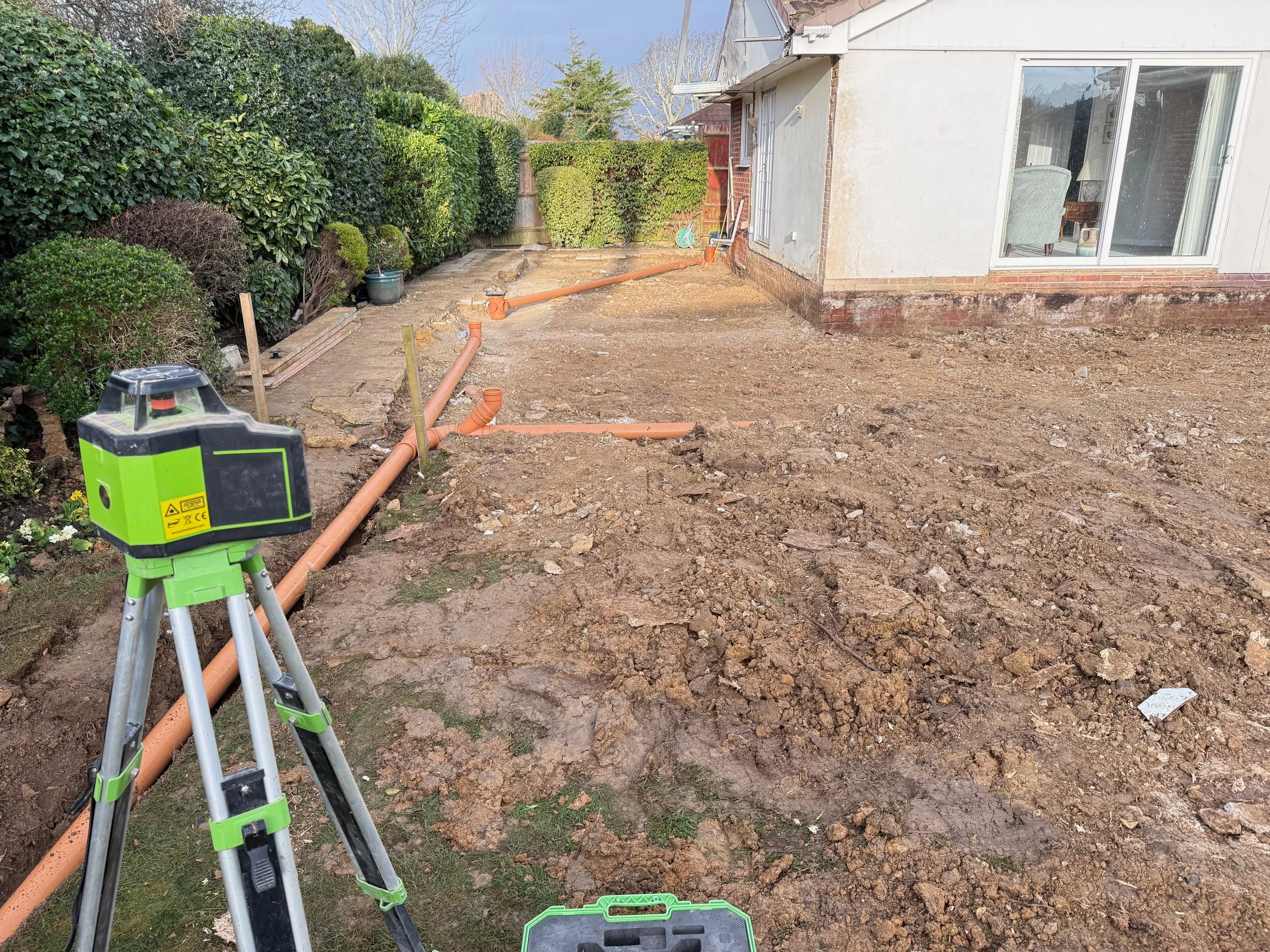 Backyard garden area under construction with exposed soil and a section of orange drainage pipes laid out inline; a green and black laser level on a tripod is in the foreground; house with sliding glass door and garden shrubs to the left.