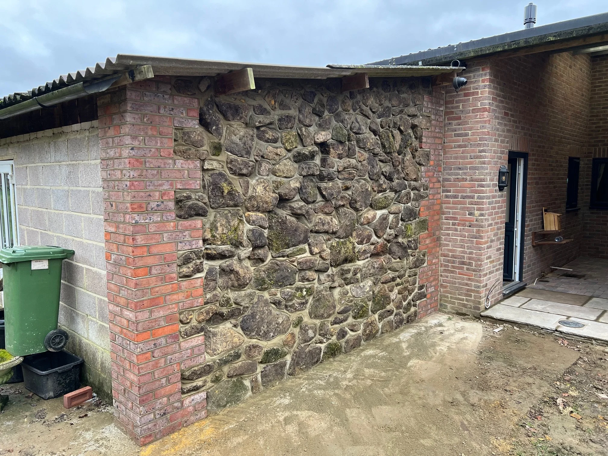 Photo of an exterior brick and stone wall of a house with a patio area and a black door. The ground is bare dirt, and there is a green trash bin to the left.