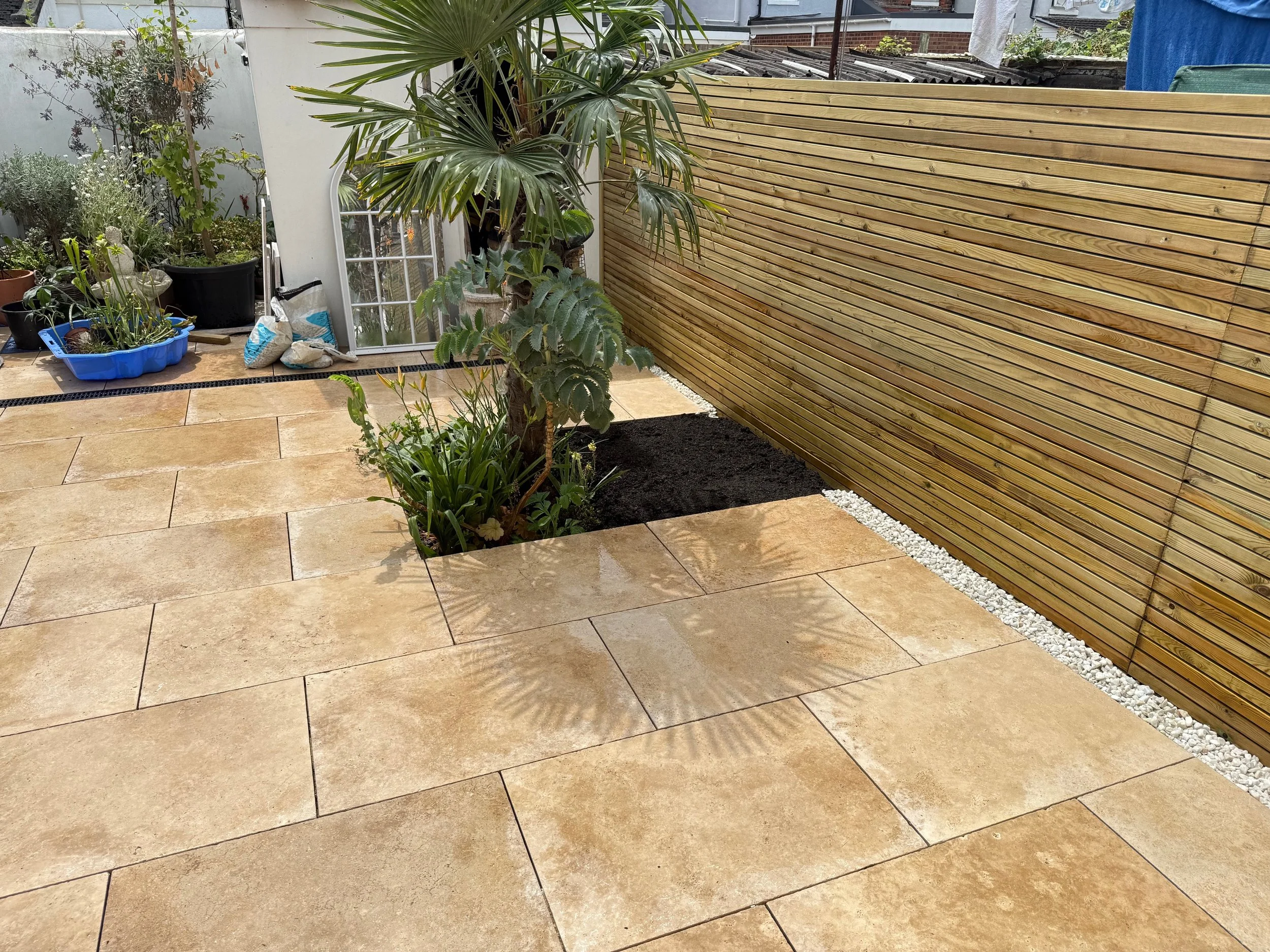 Patio area with beige stone tiles, a small tree, and various plants, enclosed by a wooden fence.