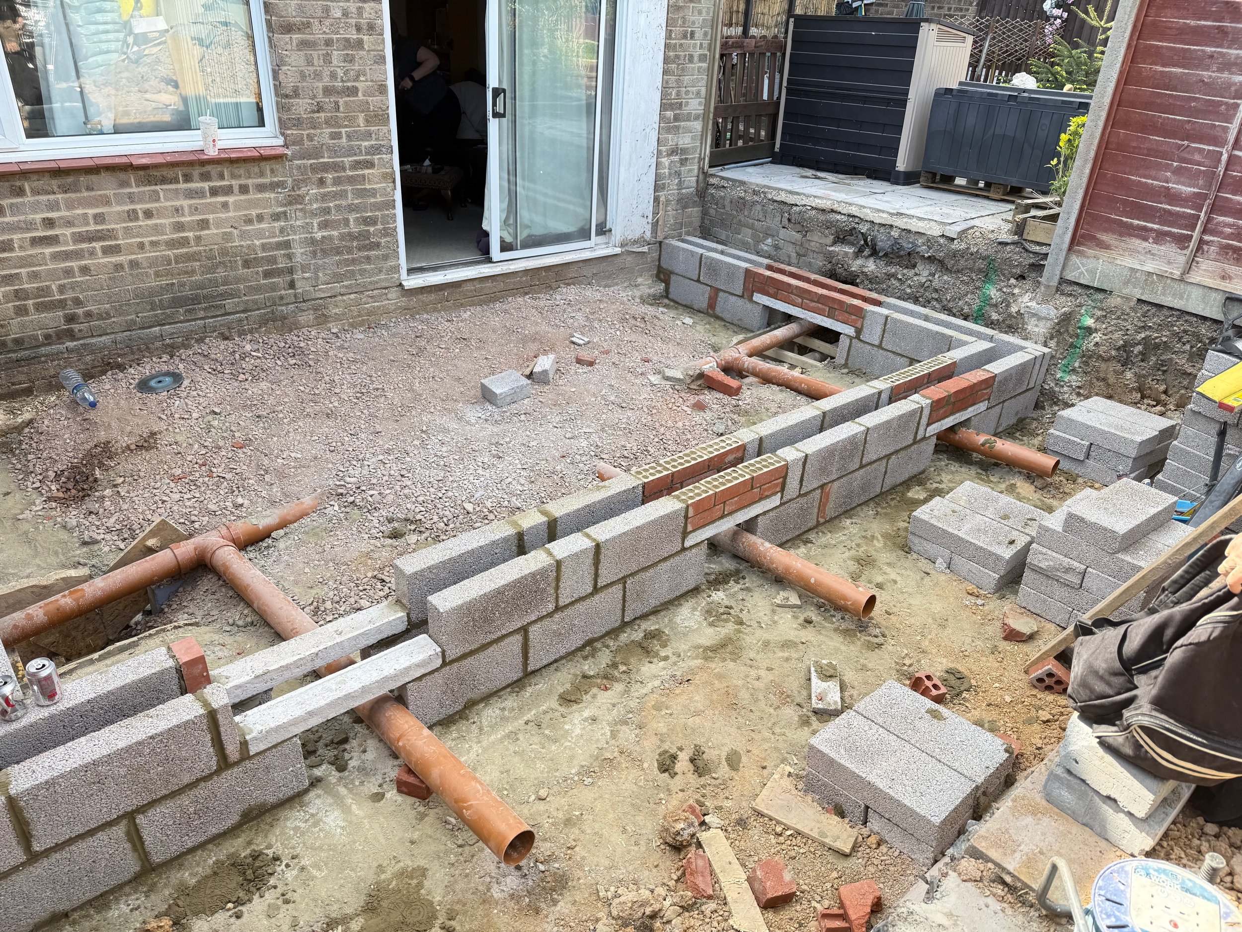 A backyard patio is under construction with cinder block and red brick walls, pipes for plumbing, and construction tools. There is a sliding glass door leading into a house and a person working inside.