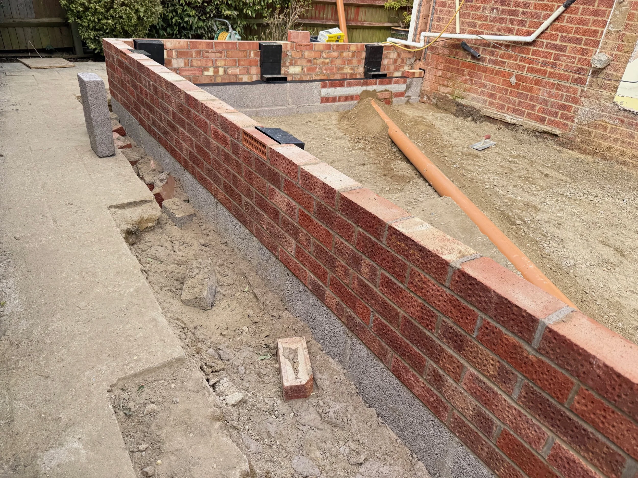 A brick and concrete wall under construction on a construction site next to a concrete sidewalk and exposed plumbing pipe.
