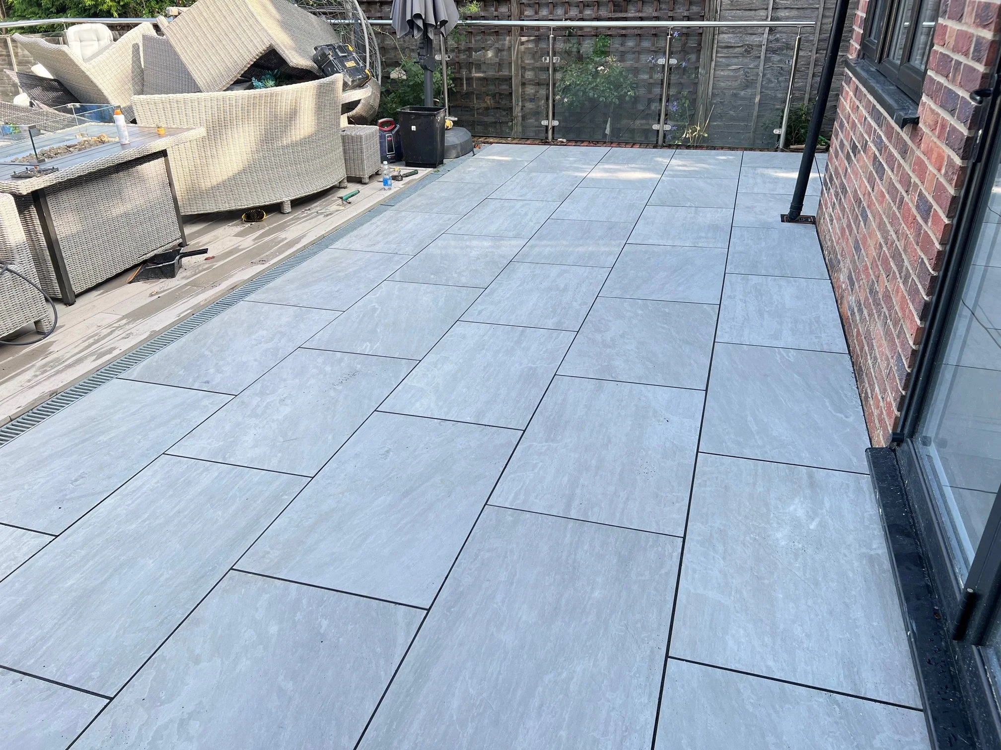 Completed light grey tiled patio with black grout lines, adjacent to a brick house with a sliding glass door, showing a cluttered area with outdoor furniture, tools, and construction materials.