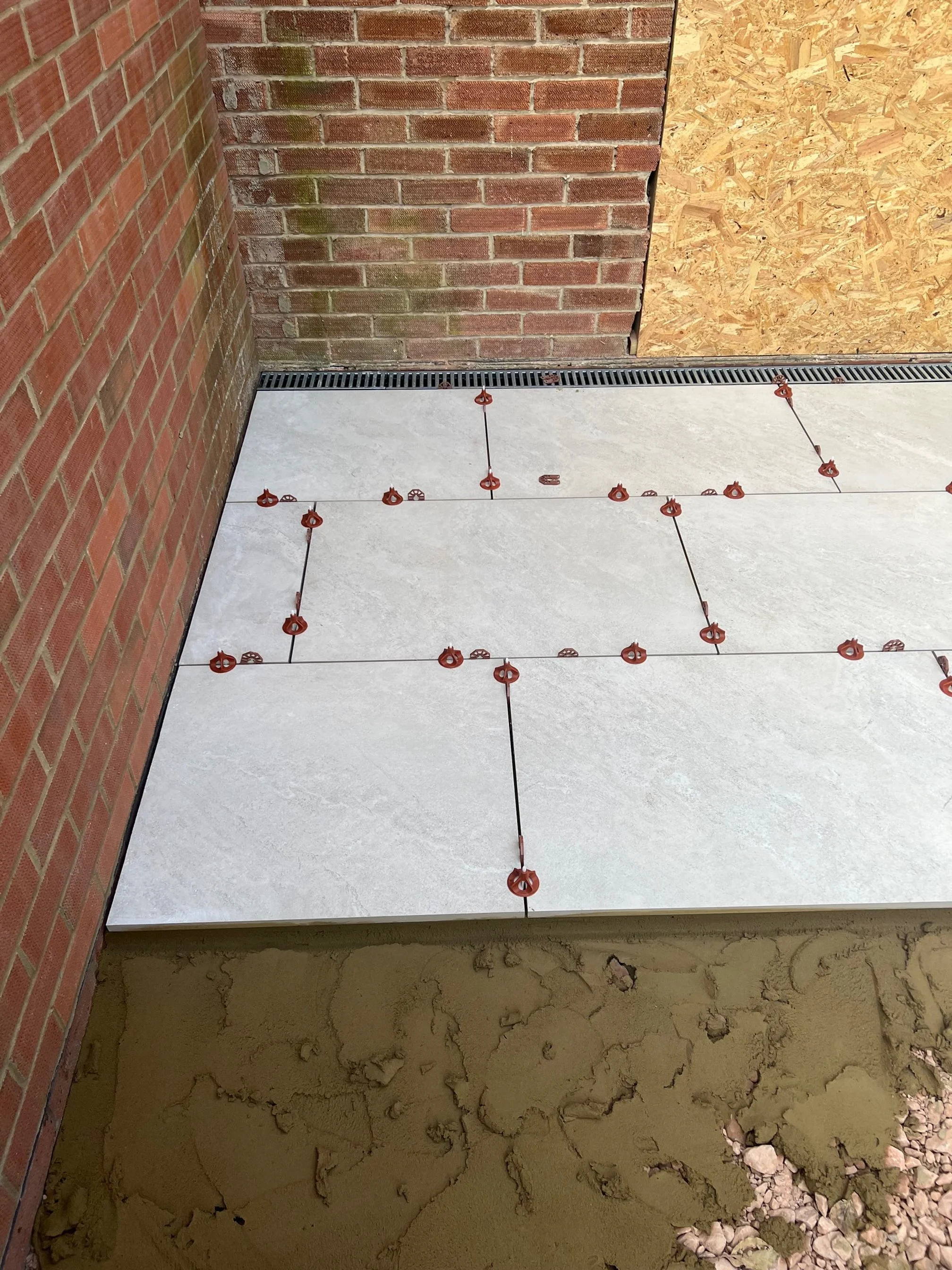 A construction site showing a partially tiled floor outside a building with brick and plywood walls. Red tile spacers are placed between large white tiles on the floor.