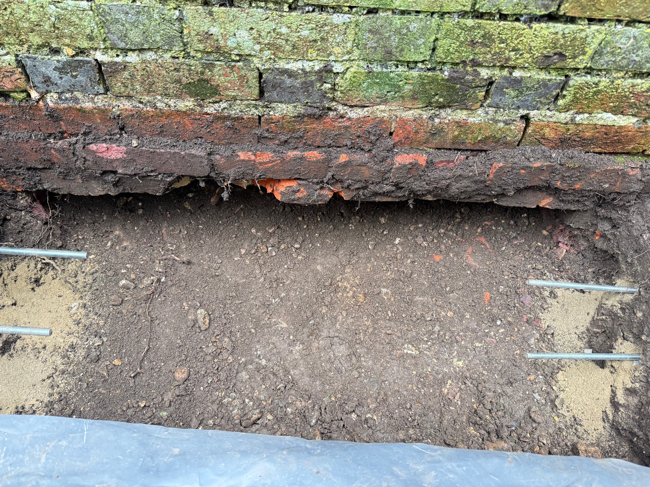 Excavation of a brick wall foundation showing dirt and metal rods for reinforcement.