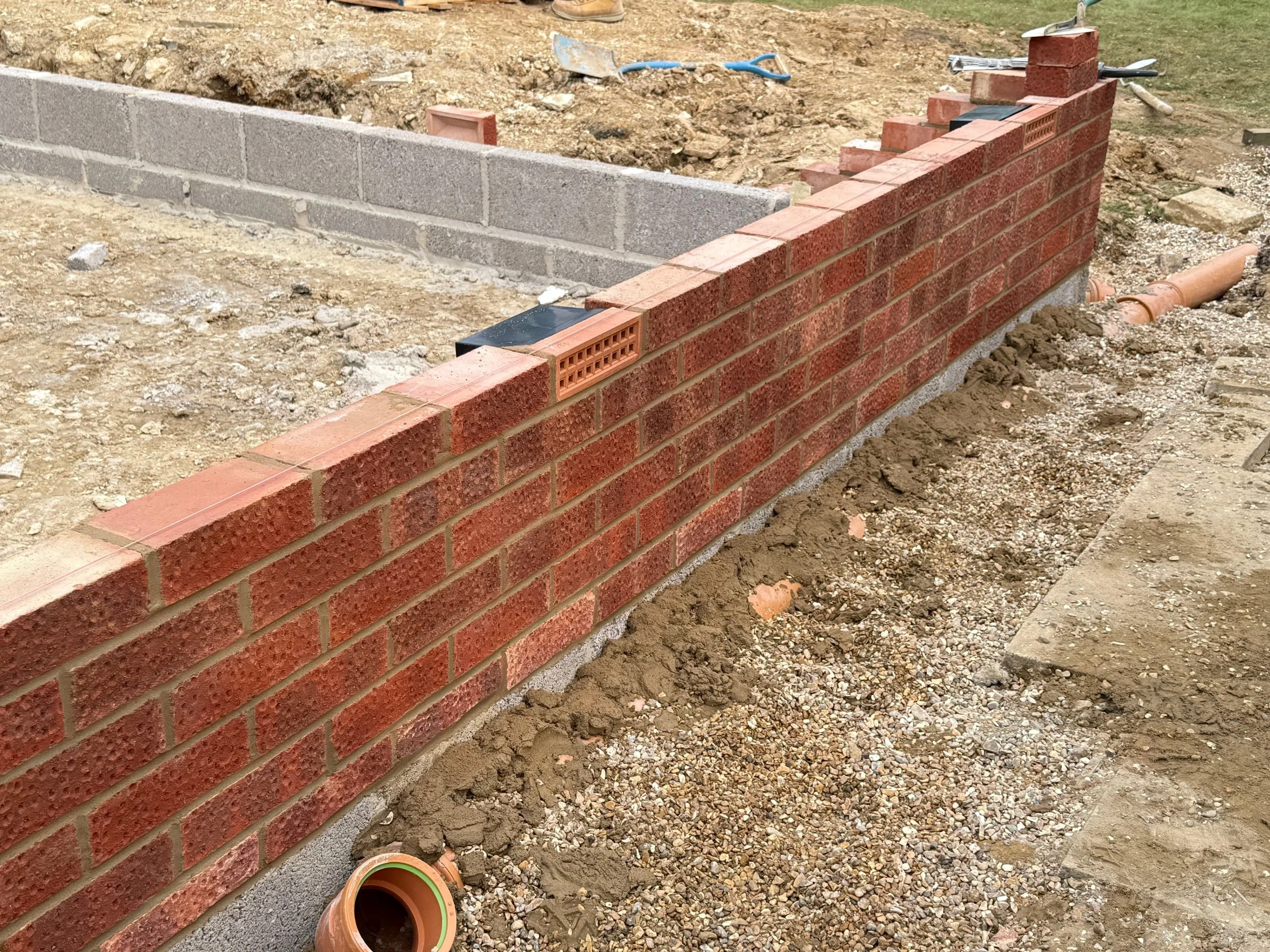 Construction site with a partially built brick wall, construction tools, and pipes on the ground.