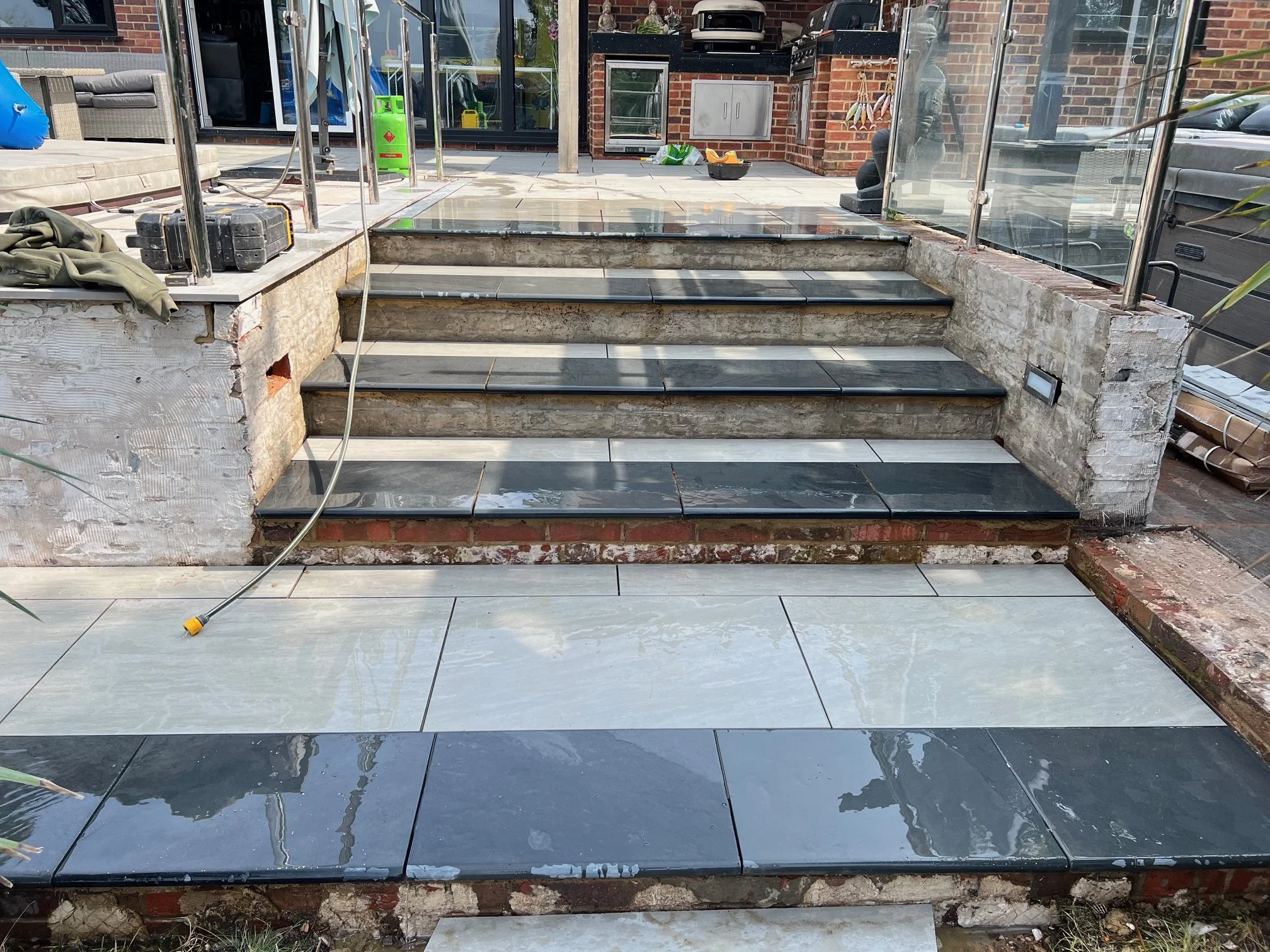 Construction of outdoor staircase with black and white tiles, surrounded by brick and concrete walls, and clear glass fencing.