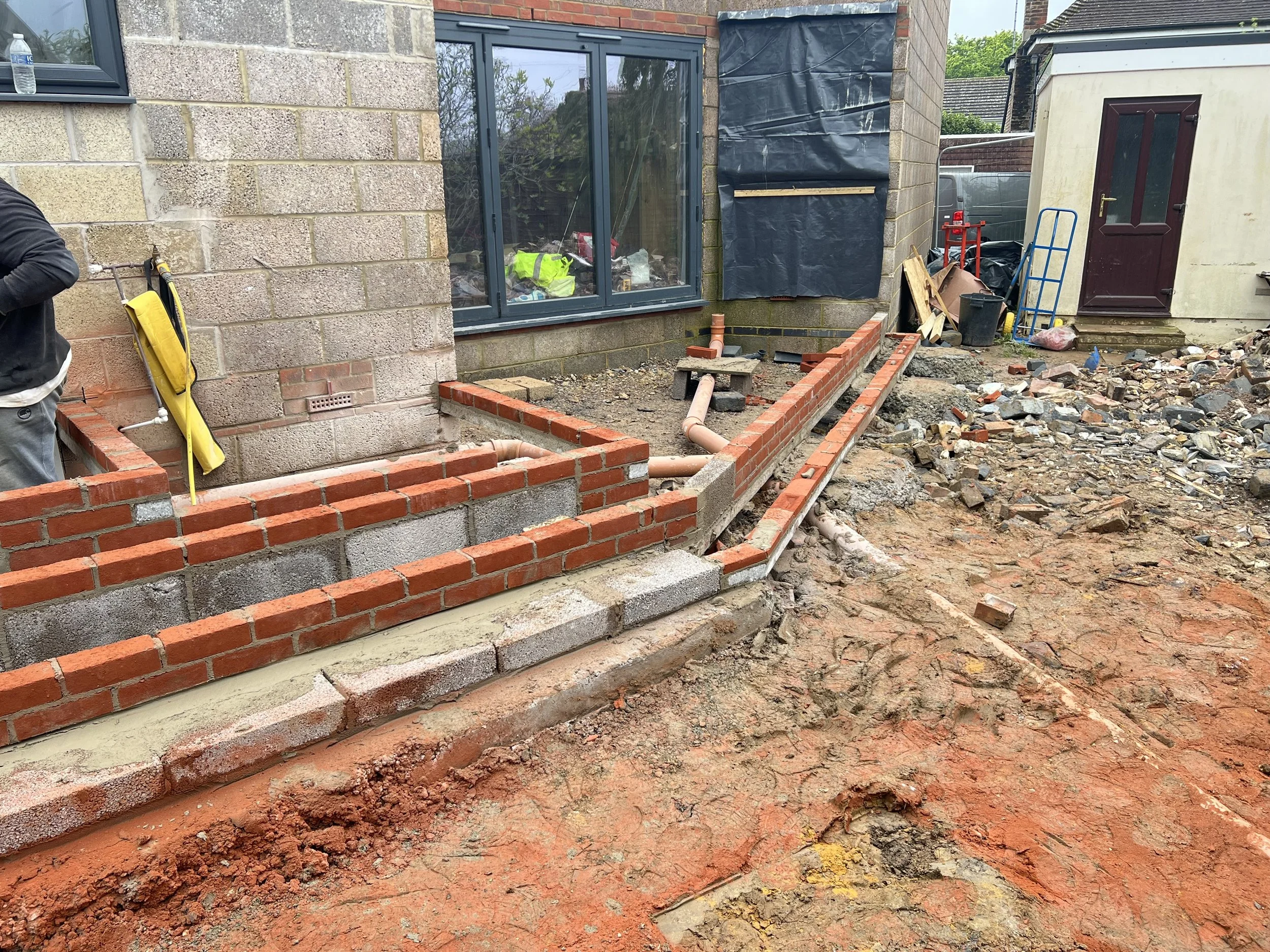 Construction site with brick walls being built around the patio area of a house, with construction tools and materials scattered around.