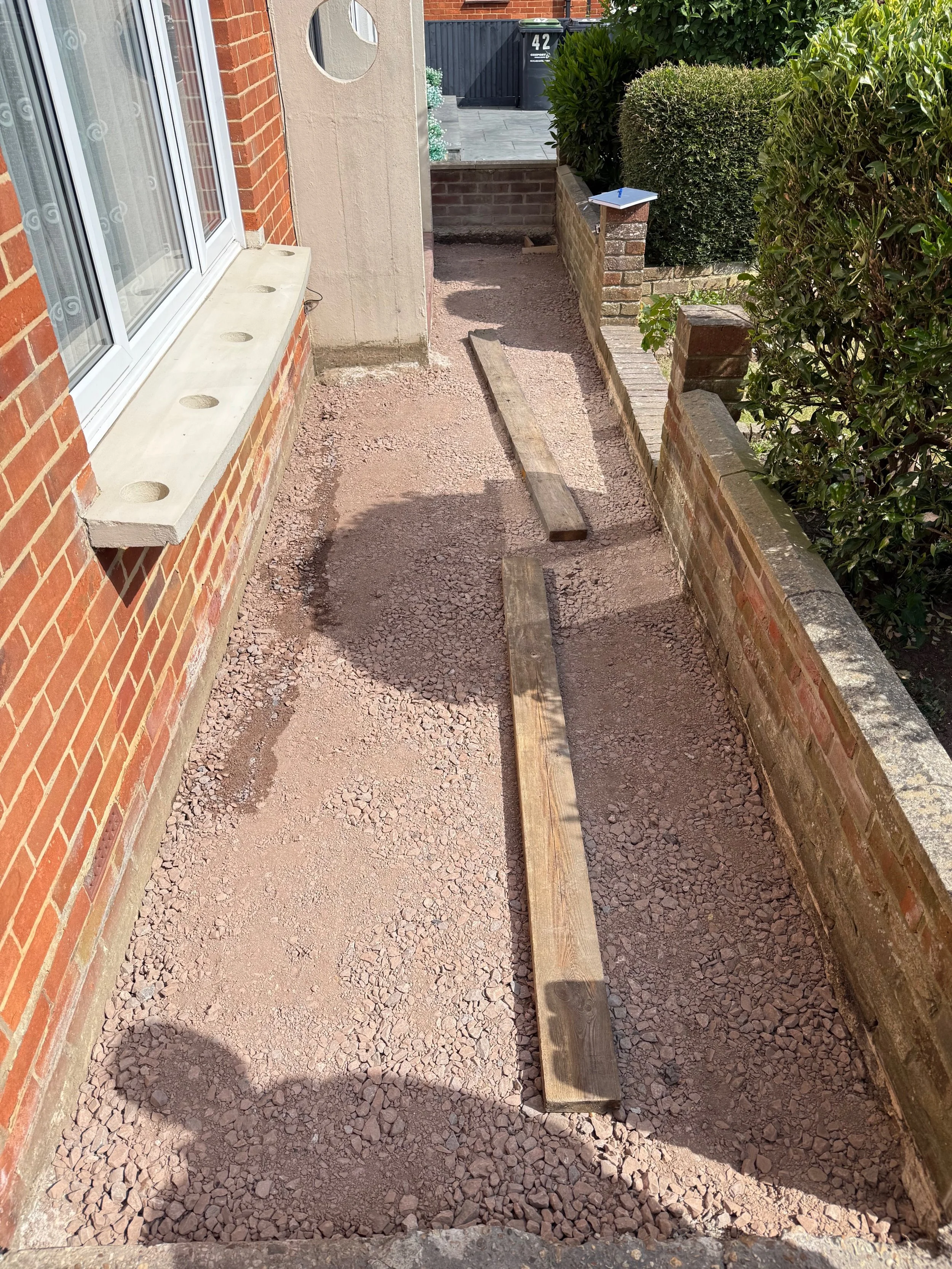 A narrow walkway under construction beside a brick house with a large window. Two wooden planks are laid on the ground, and a small brick wall and various bushes are visible along the path.