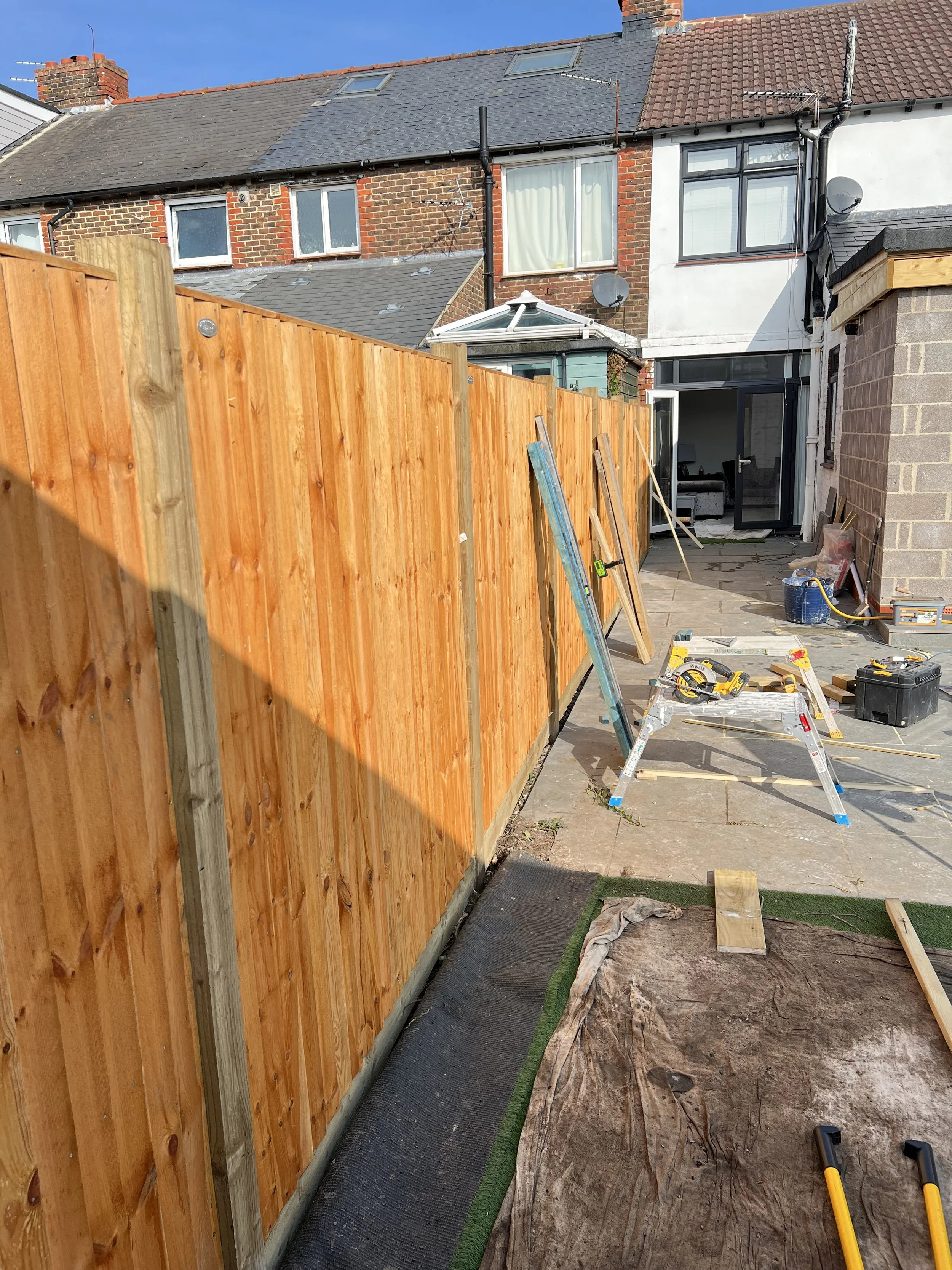 A backyard area under construction with a new wooden fence being installed. Tools, including a saw, loppers, and a ladder, are scattered around. The house with brick and white siding, neighboring houses, and a clear blue sky are visible in the backgr