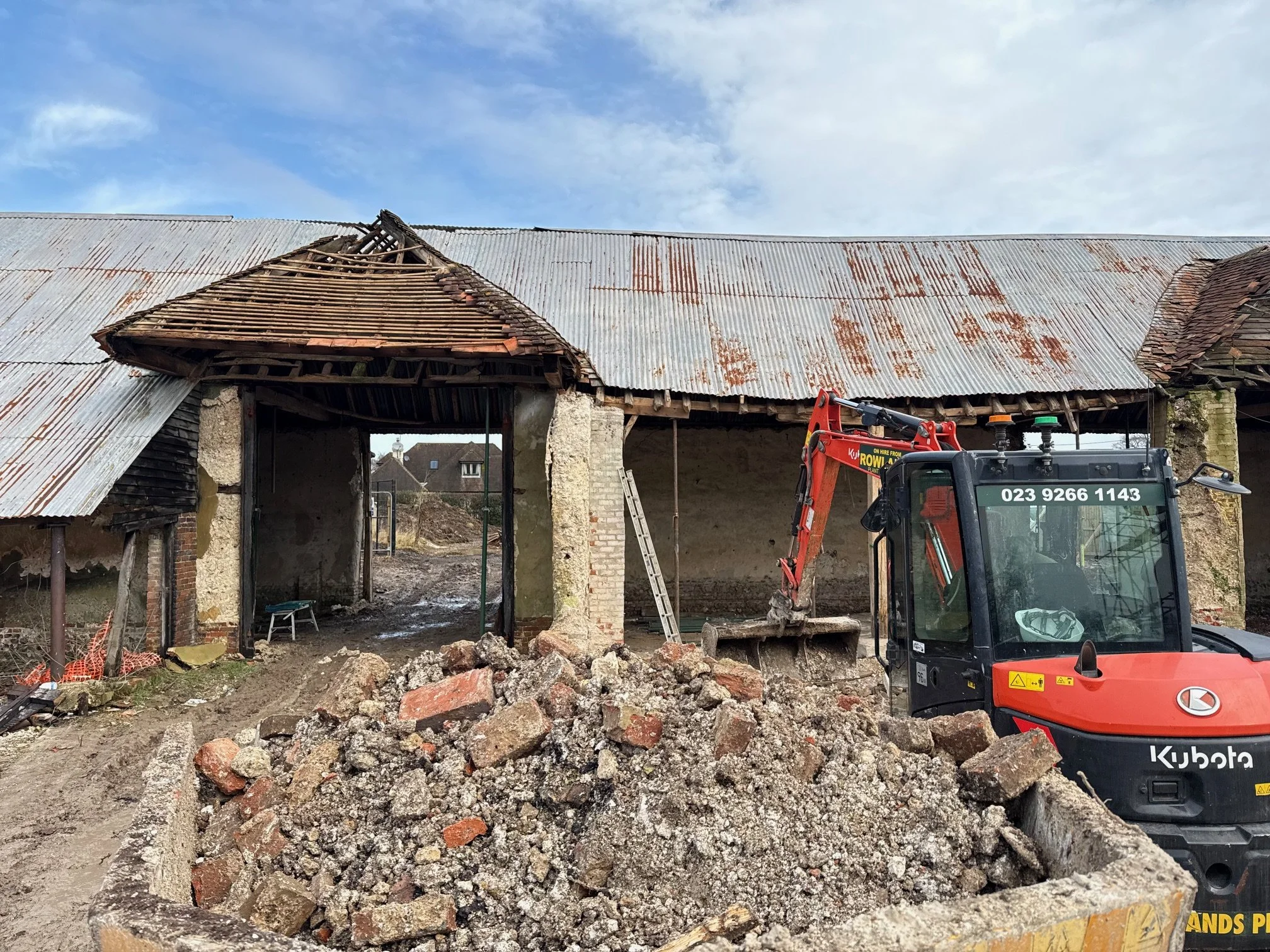 Construction site with a bulldozer demolishing an old, damaged roof of a building, with debris and bricks in the foreground, under a partly cloudy sky.