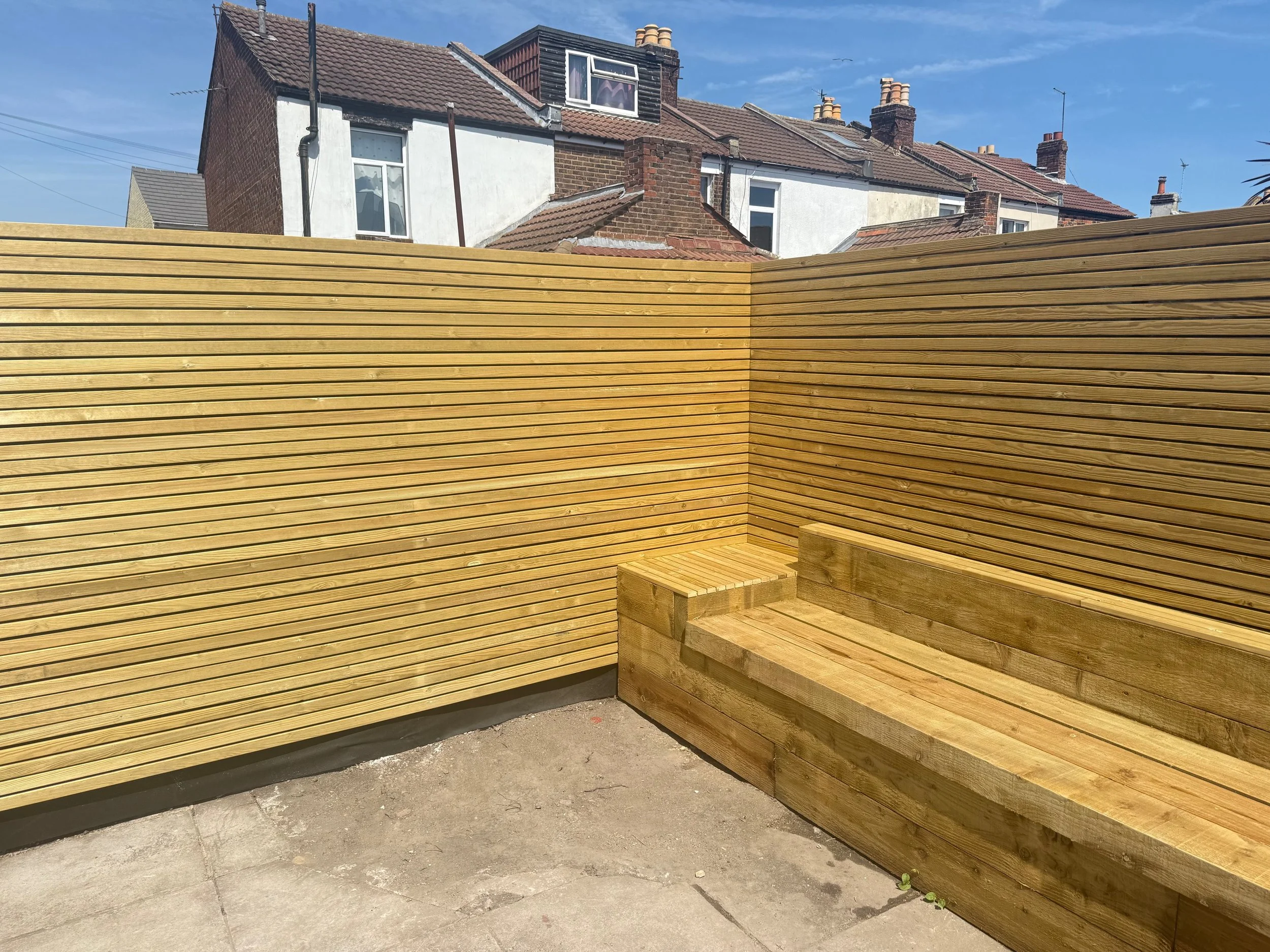 Wooden outdoor bench with backrest in a small enclosed yard, surrounded by wooden fencing, with neighboring houses visible in the background under a clear blue sky.