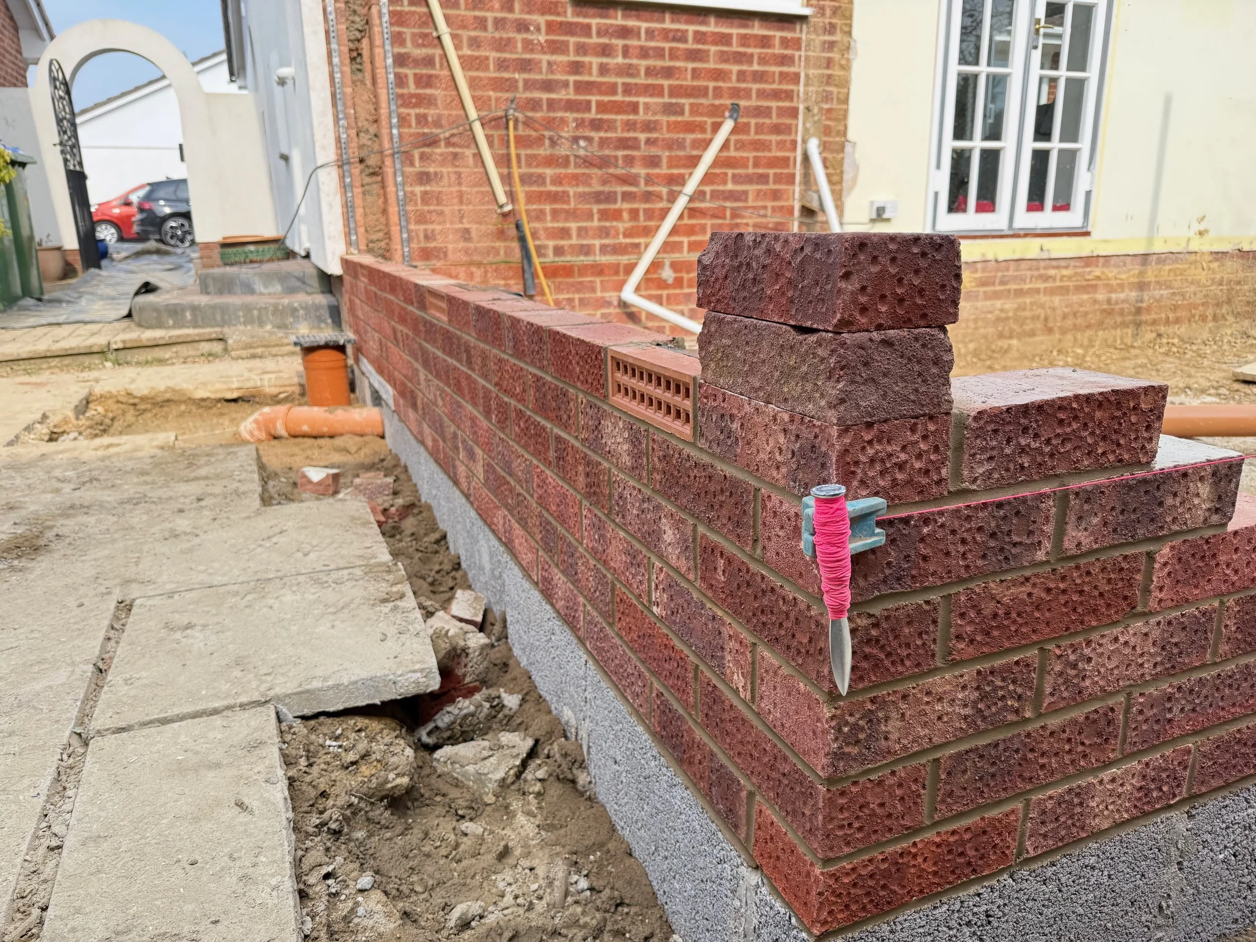 A brick wall under construction with three bricks placed on top, a pink string for alignment, and construction tools in the background.