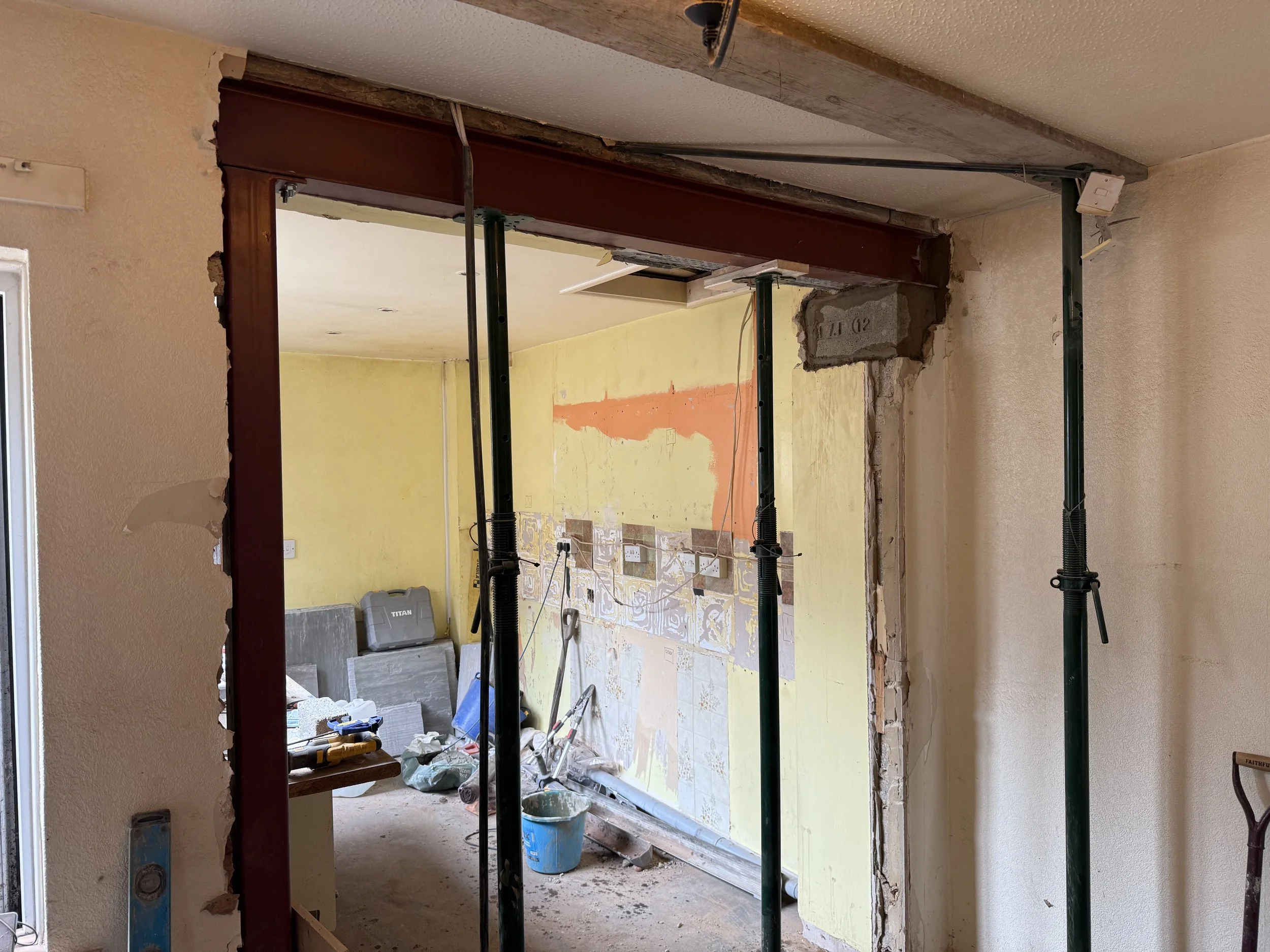 A room under renovation with a partially removed wall, exposed beams and drywall, construction tools, and equipment scattered on the floor.