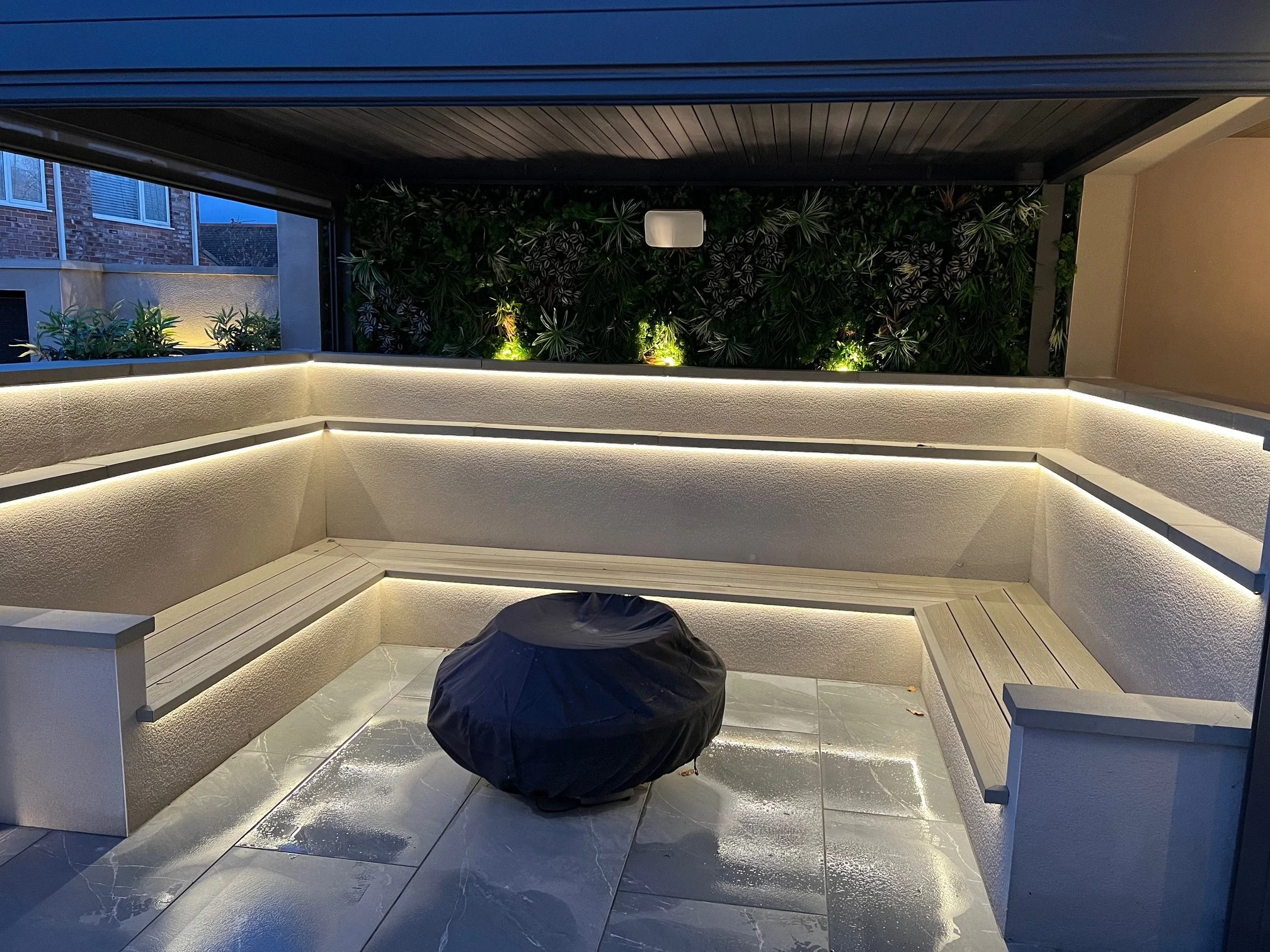 Modern outdoor patio with built-in bench seating, illuminated with LED strip lighting, and a covered grill in the center, overlooking a green vertical garden at dusk.