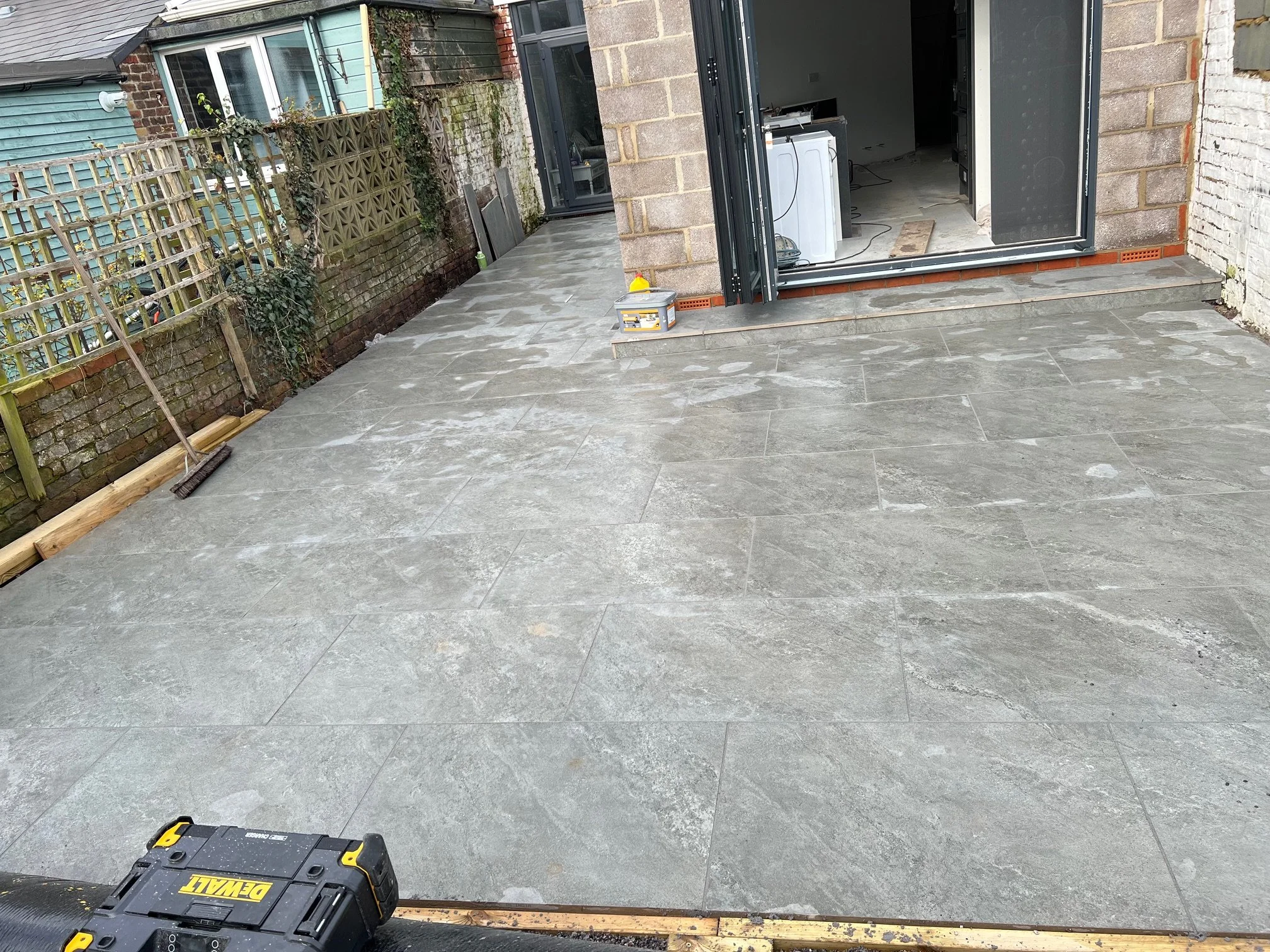 Newly installed gray stone patio outside a house under construction, with construction tools and materials visible, and an open doorway leading inside.