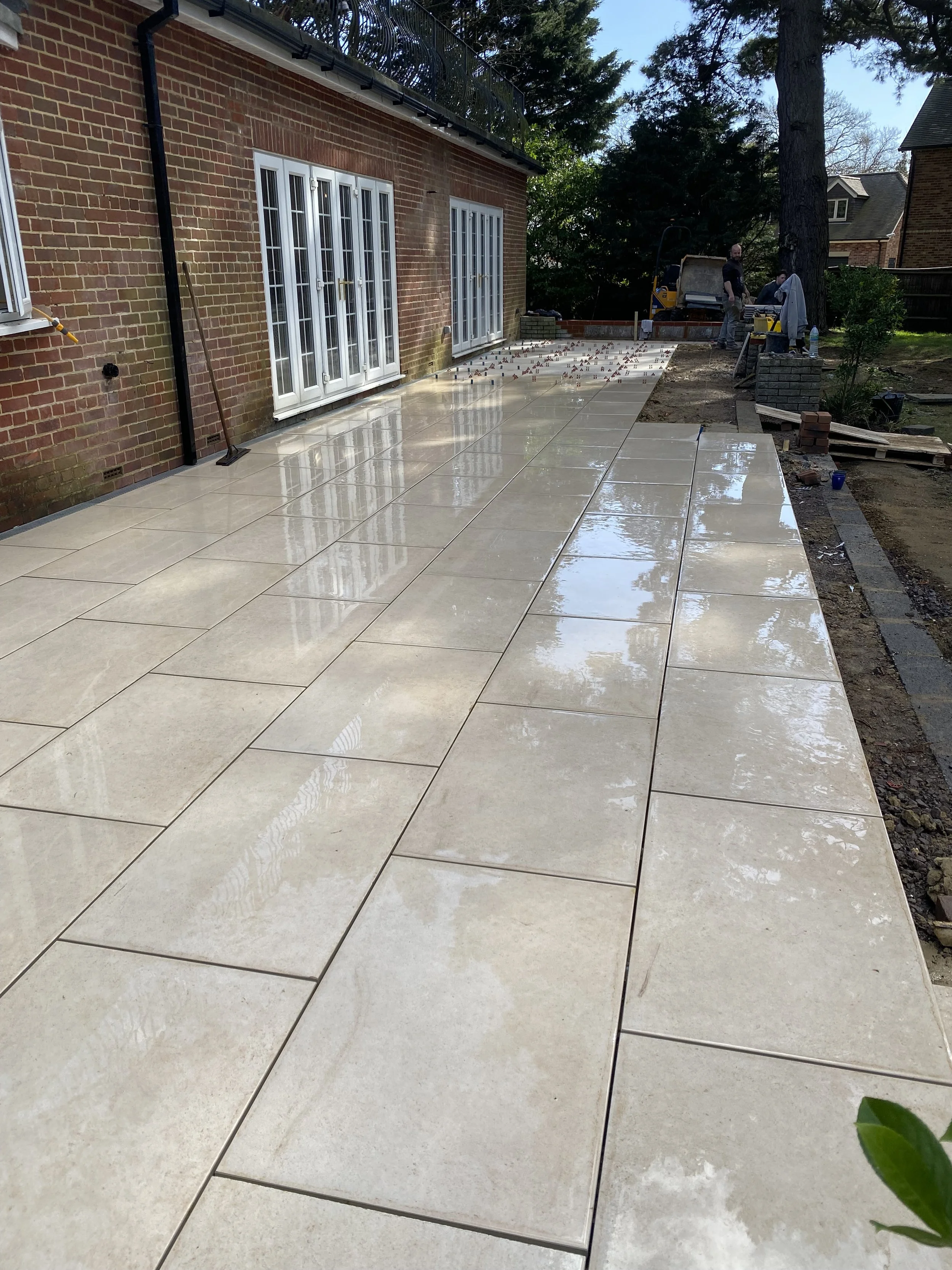 A recently constructed patio with glossy, beige tiles outside a brick house, with workers and equipment visible in the background during daylight.