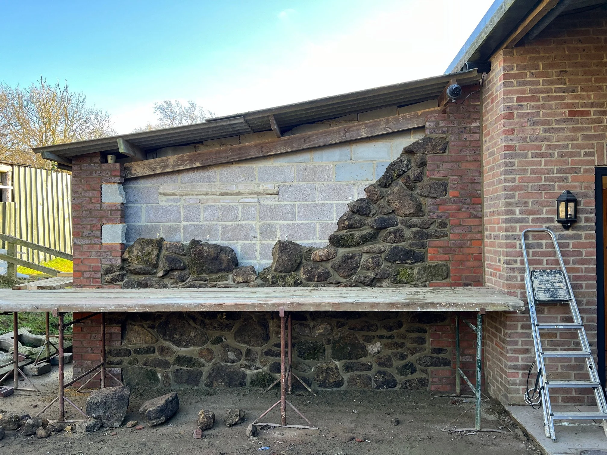 Resurfacing of an outdoor brick wall with concrete blocks and large rocks, with a scaffold and ladder nearby during construction.