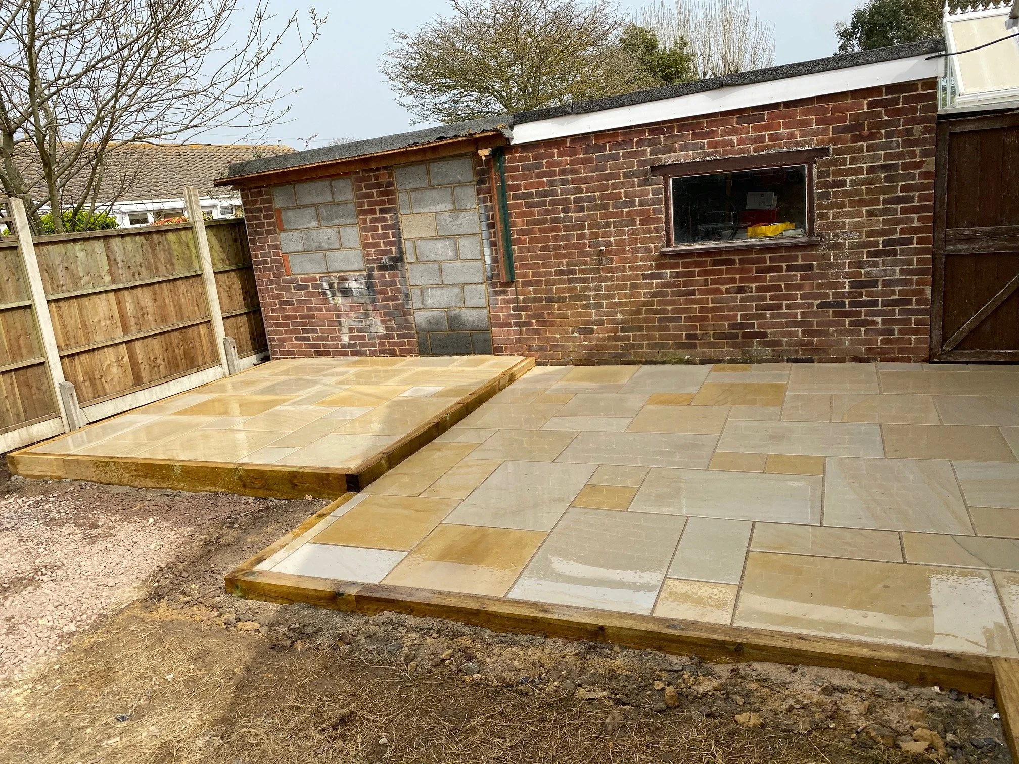 Newly paved patio with beige tiles in front of a brick shed, surrounded by a wooden fence, with a small window on the shed and some bare trees in the background.