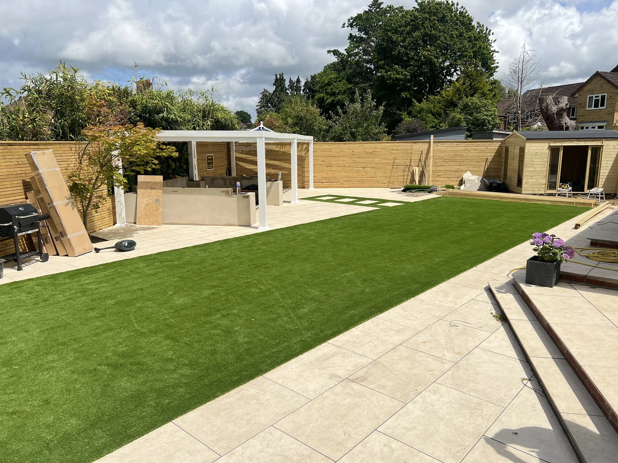 Backyard with a green artificial lawn, beige patio tiles, a wooden fence, and a small wooden outbuilding. There is a covered outdoor seating area in the corner, and various construction materials and tools are scattered around, indicating ongoing wor