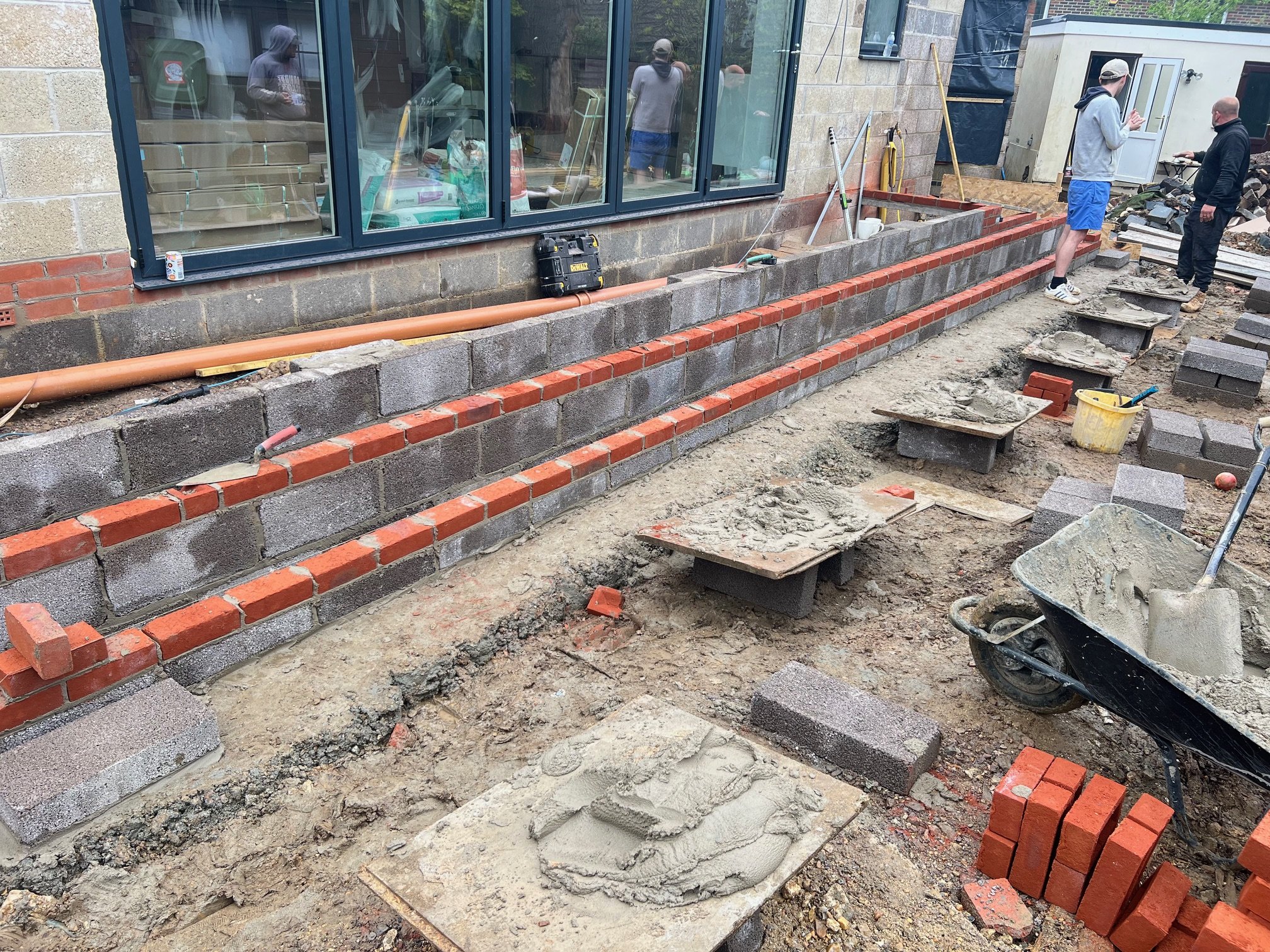 Construction workers building a brick and cinder block retaining wall outside a house, with tools and materials scattered around.