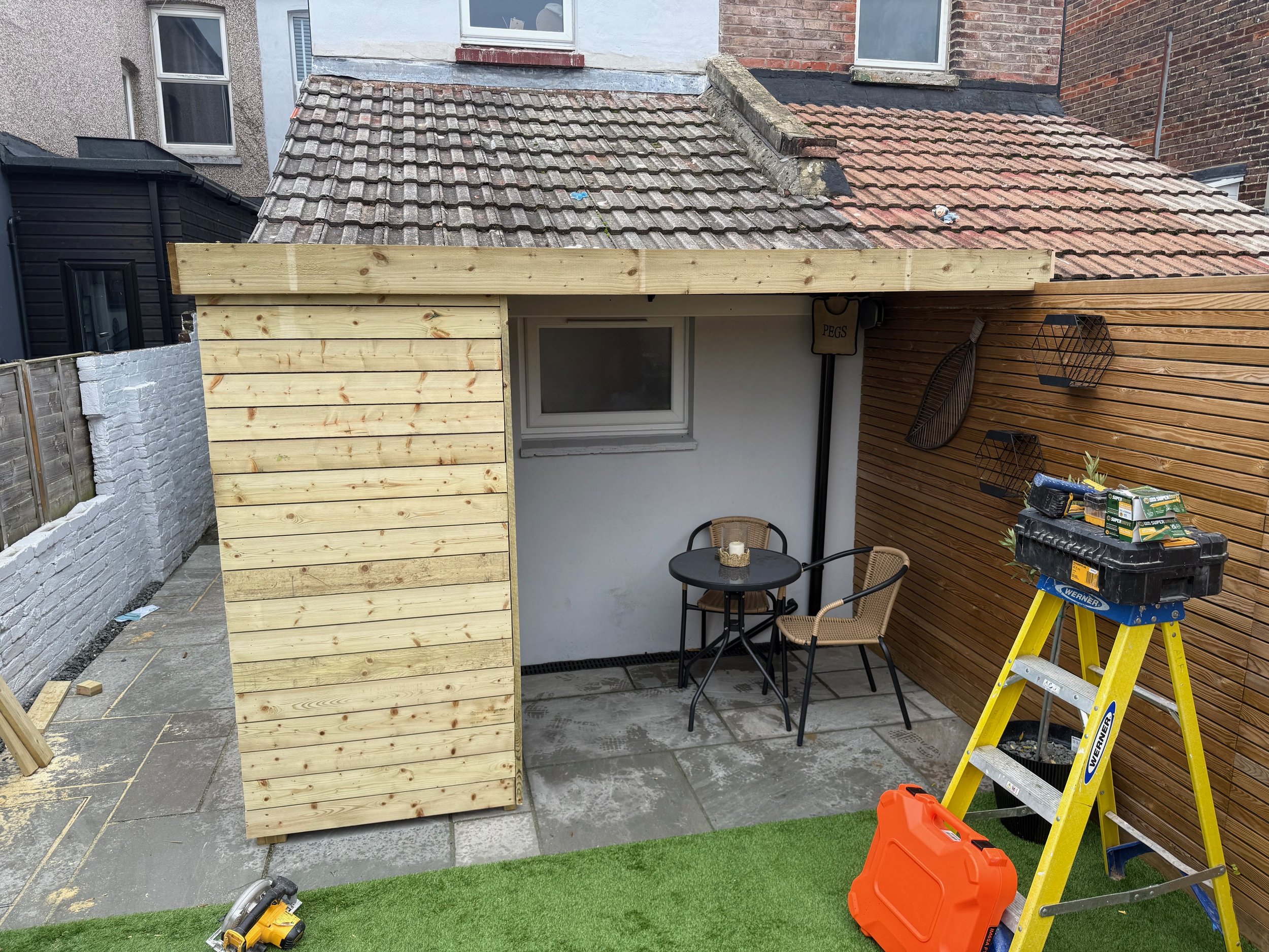 A backyard patio area with a small seating table and two chairs, partially enclosed with wooden panels, construction tools, and materials present.
