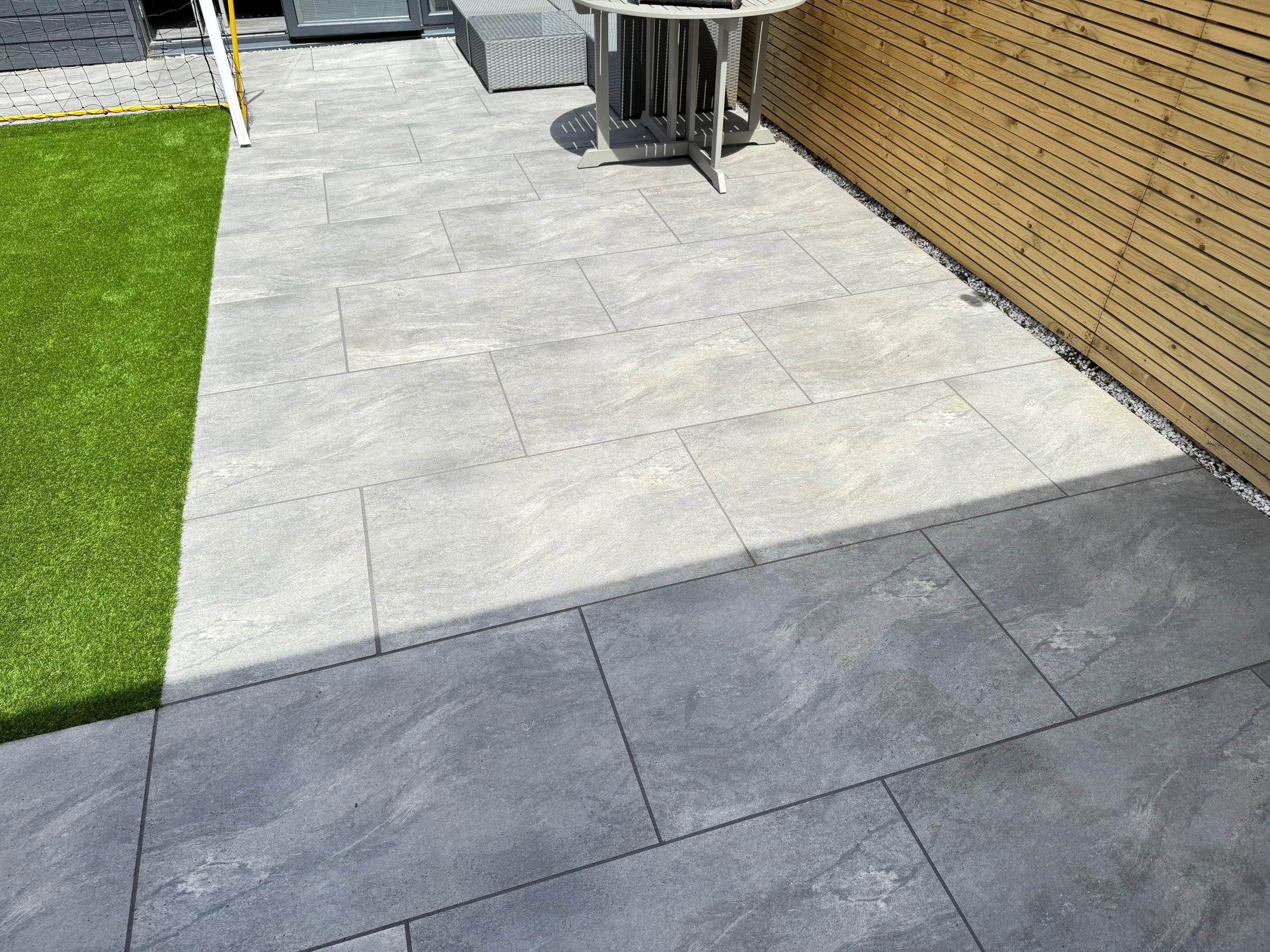 Outdoor patio with large gray tiles, a small patch of artificial grass, a wooden fence, a sports goal, and a metal outdoor furniture set.