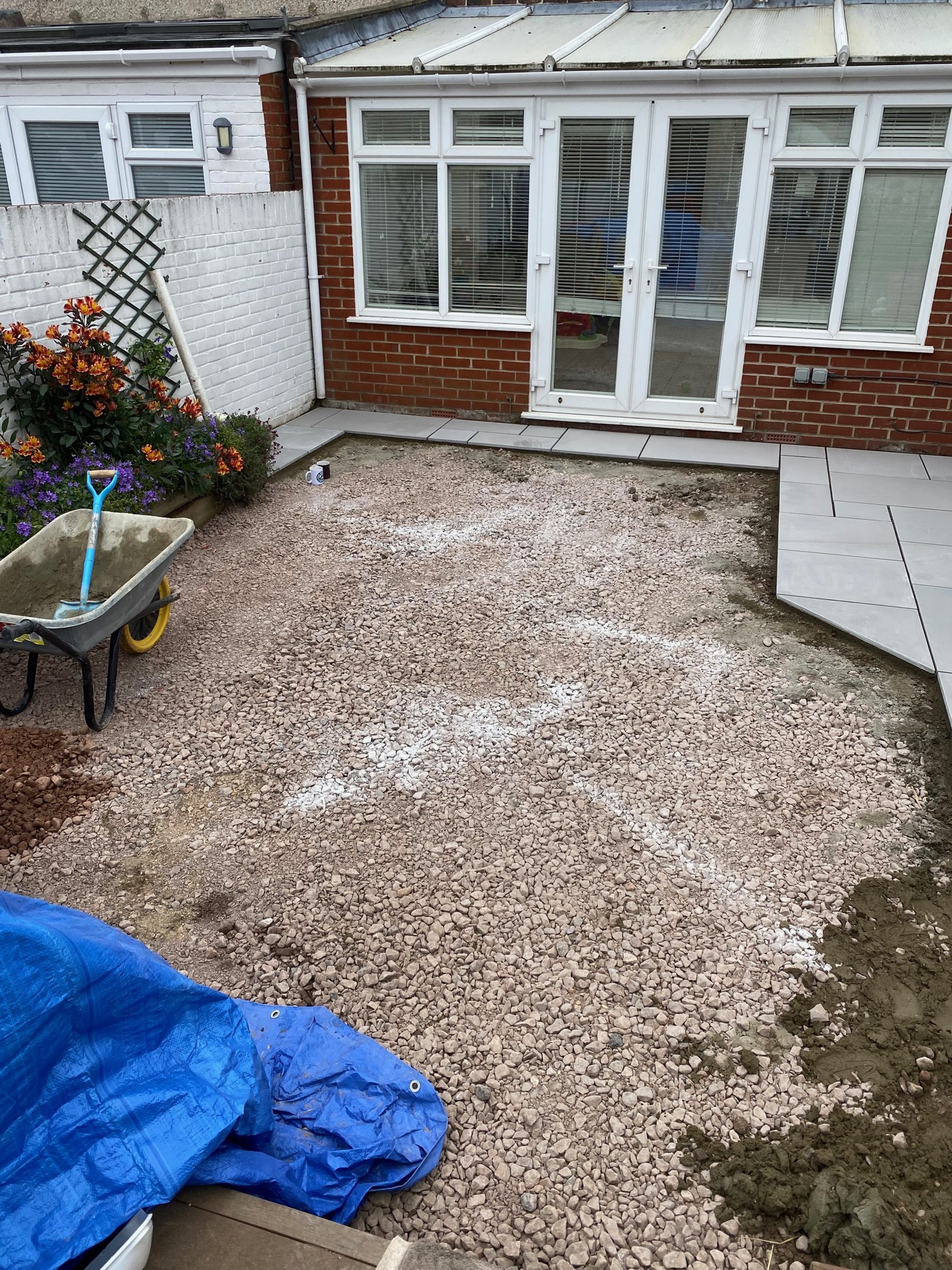 Backyard patio under construction with gravel, a wheelbarrow, and partially laid concrete tiles. A white brick wall with flowers and a white fence are visible, along with gray paving stones near the house.