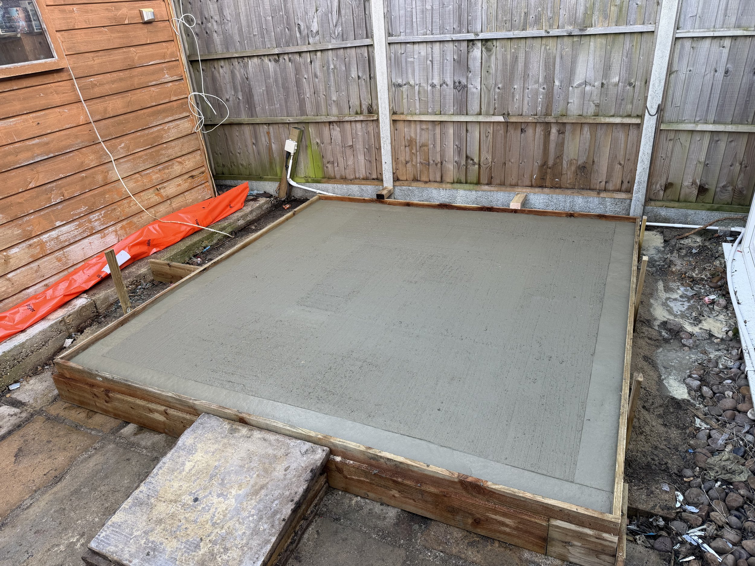 Freshly poured concrete slab in a backyard, surrounded by wooden framing, with a concrete step at the front.