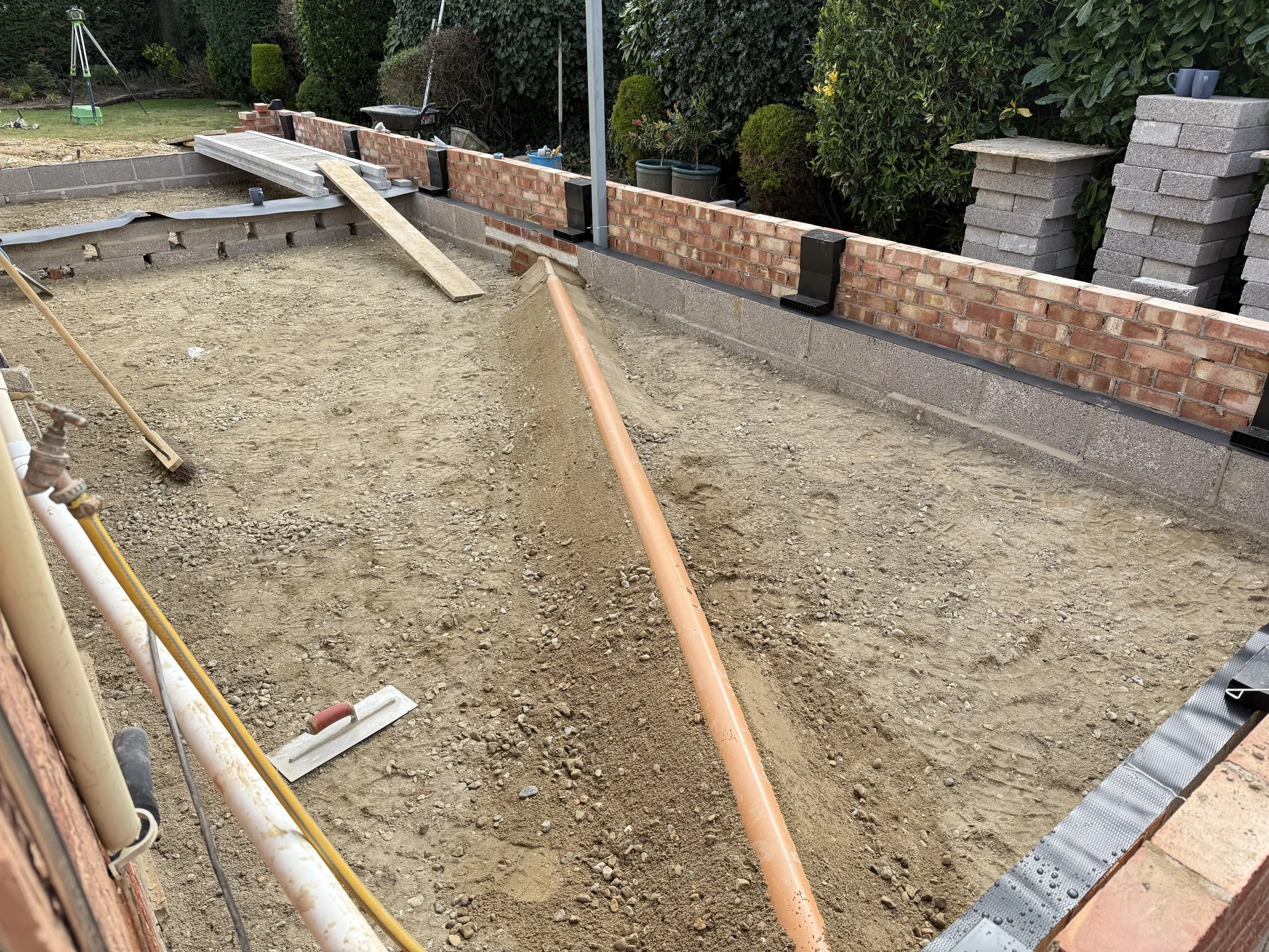 Construction site with a partially built brick and concrete block wall, open dirt ground, construction tools, and building materials.