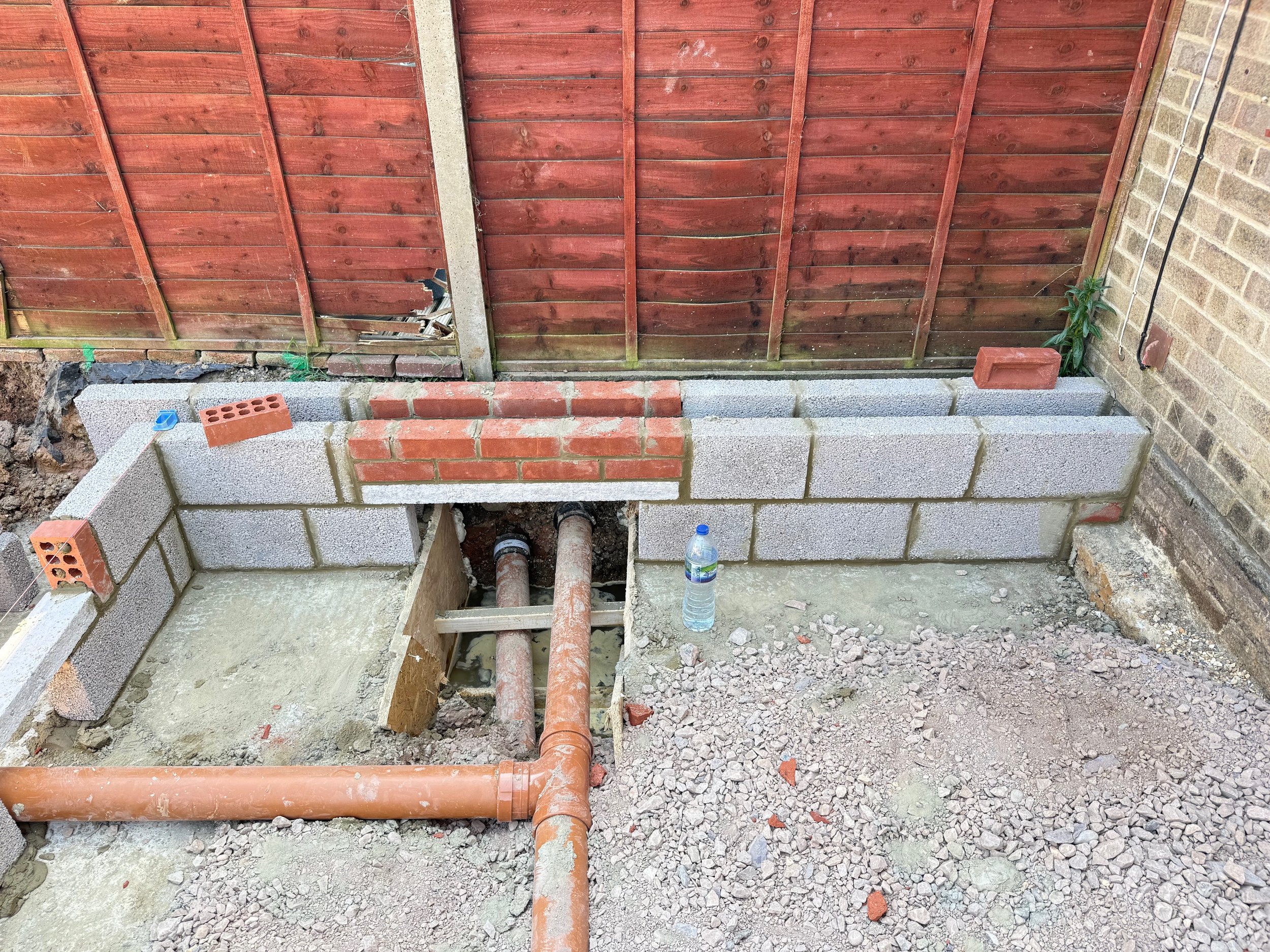 Construction site with cinder block walls, exposed pipes, and dirt ground, next to a wooden and brick fence.