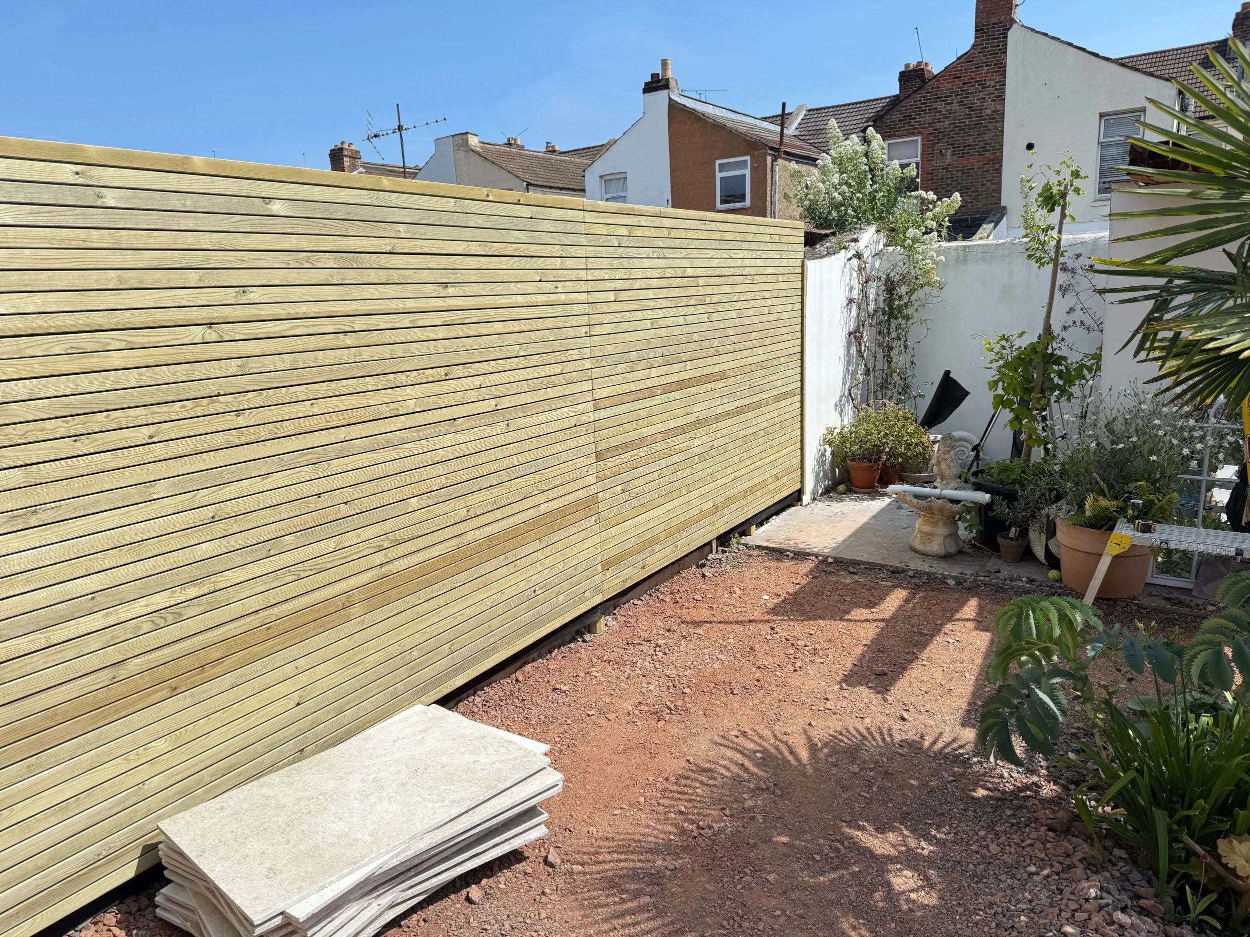 Side view of a backyard with a newly installed wooden fence, potted plants, and construction materials on the ground.