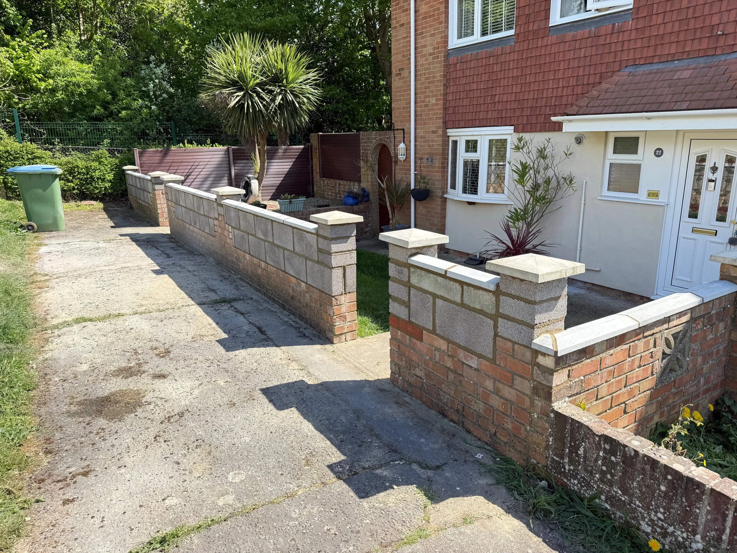 Front view of a residential house with a low brick and concrete block wall, a driveway, and garden plants, including a palm tree.