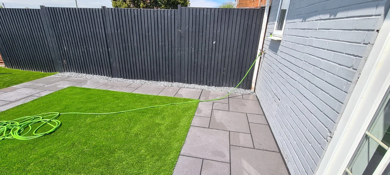 Side yard with gray concrete pavers, green artificial grass, white brick house wall, black wooden fence, and garden hose connected to a water source.