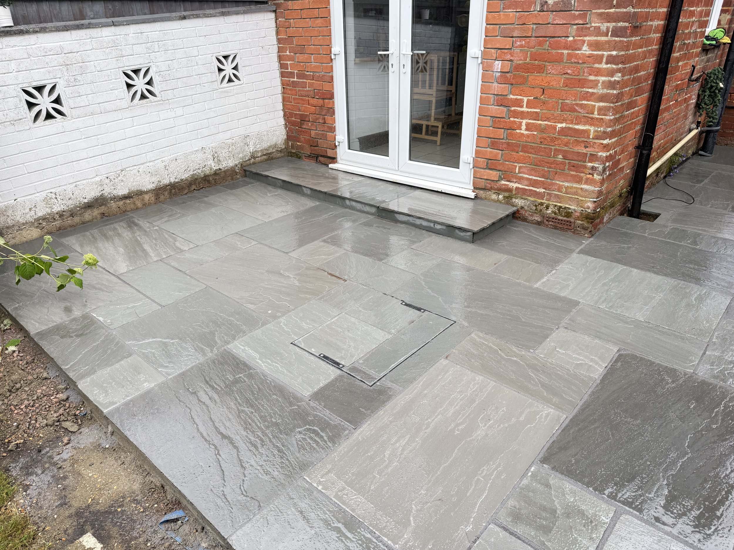 Recently paved outdoor patio area with grey stone tiles and a small step leading to a white door with glass panes, adjacent to a brick wall and a white painted wall.