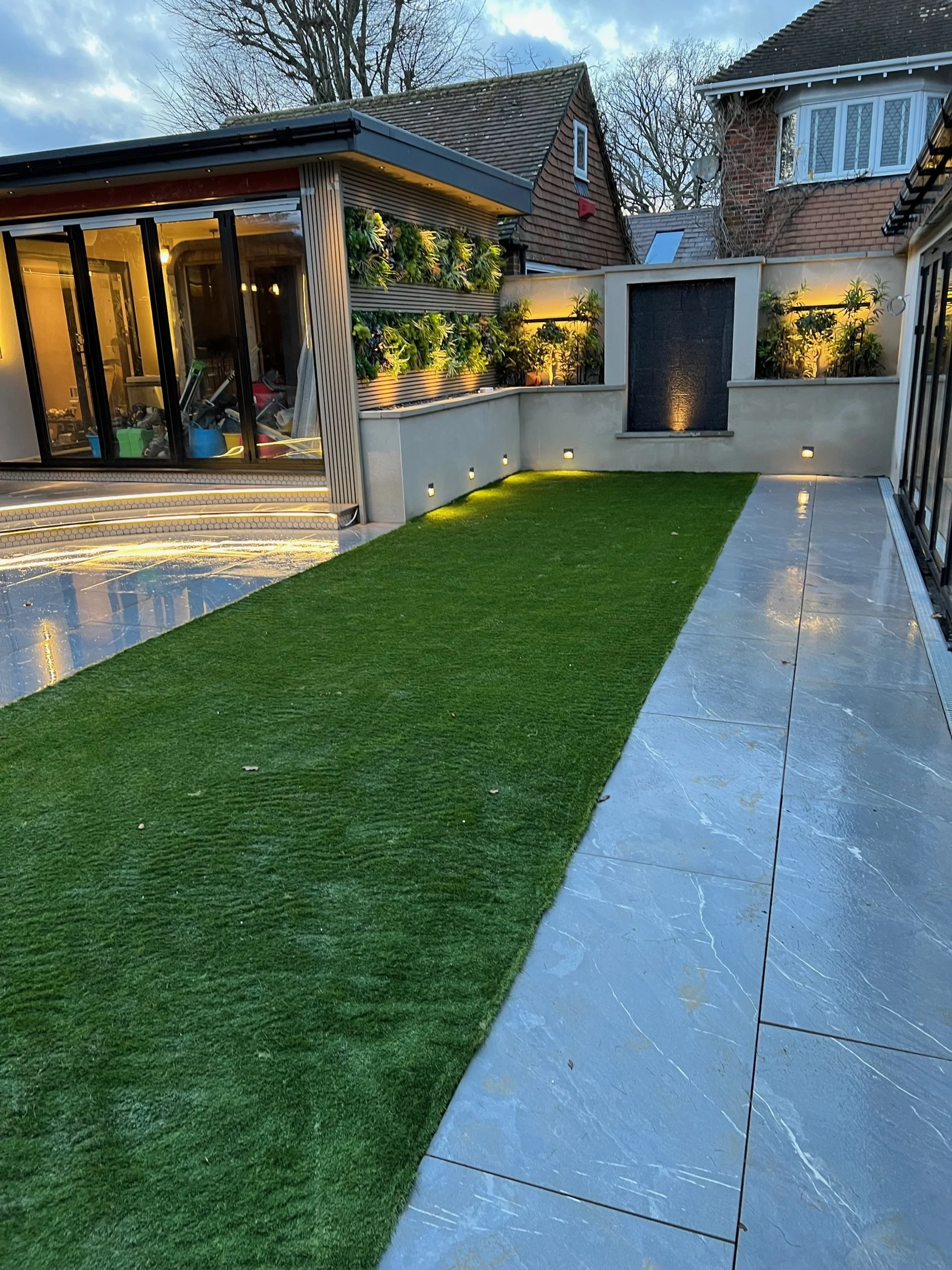 Backyard patio with artificial grass, marble tiles, a water feature with a fountain, and a garden wall with potted plants and lighting.