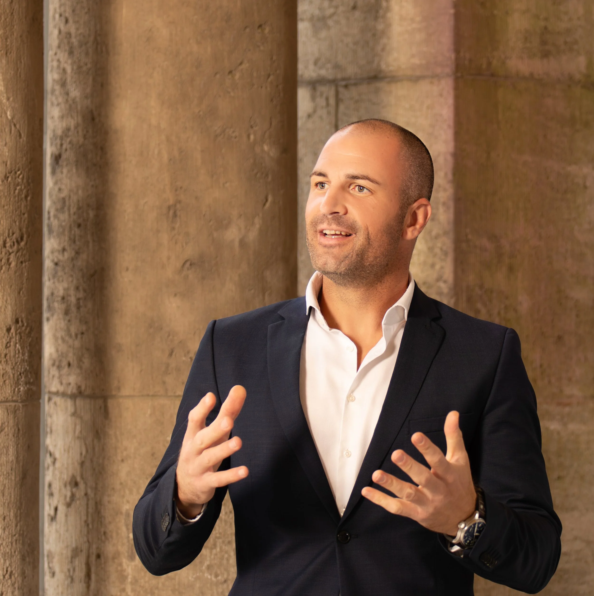 A man in a formal suit and white shirt gesturing while speaking in front of stone columns.