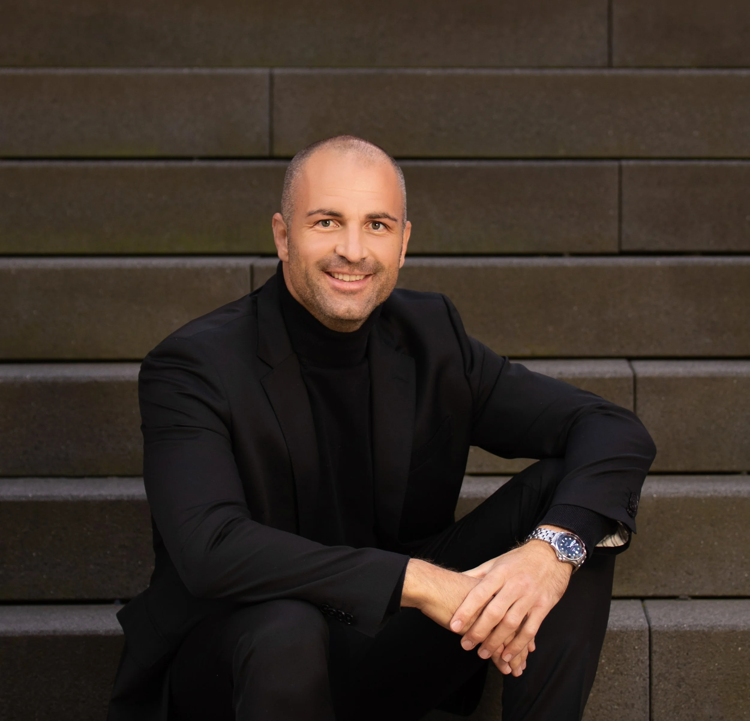 A man with a shaved head and beard wearing a black suit and turtleneck, sitting on brown stairs, smiling at the camera, with a silver watch on his left wrist.