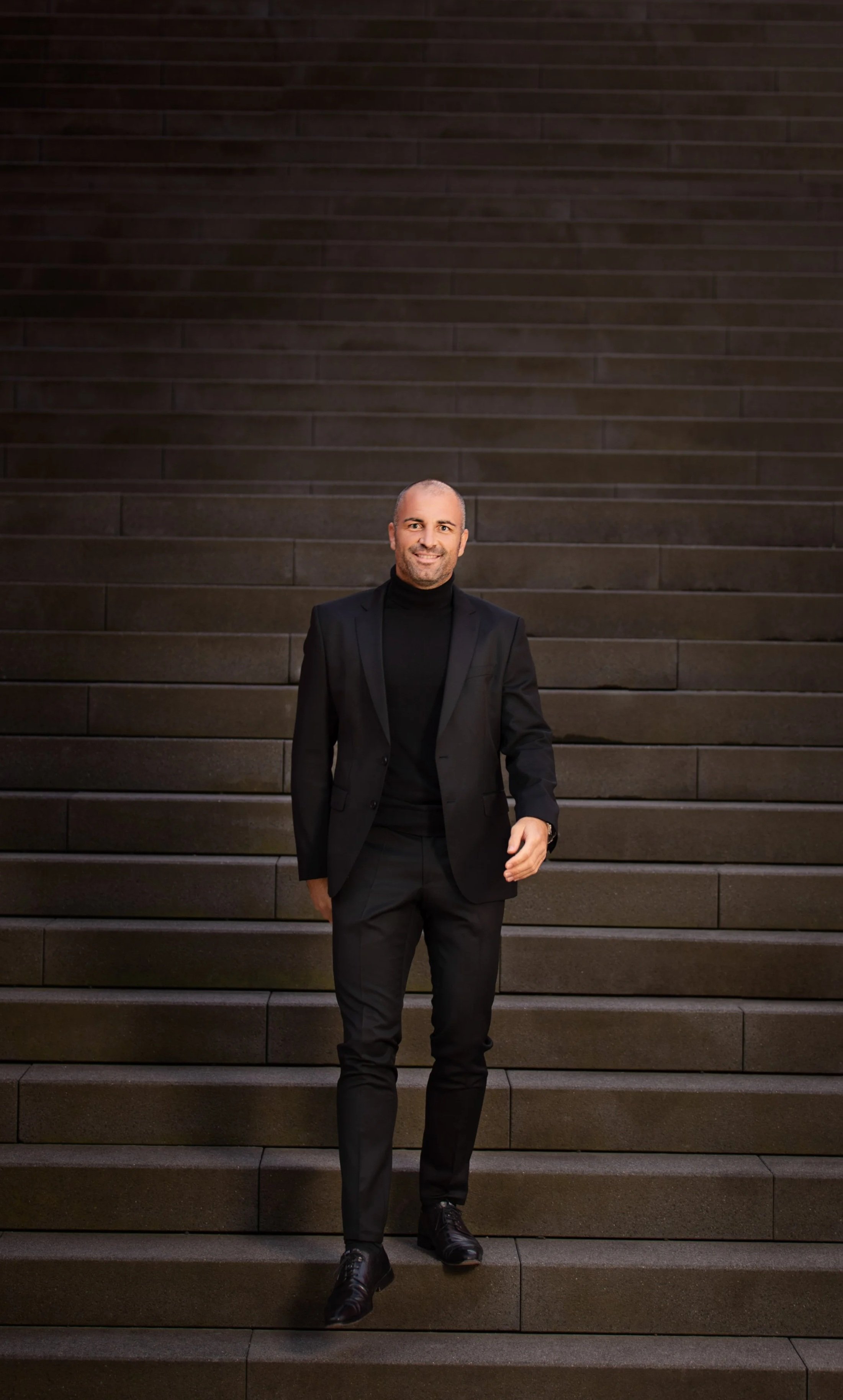 A man in a black suit and turtleneck walking down a dark-colored staircase.