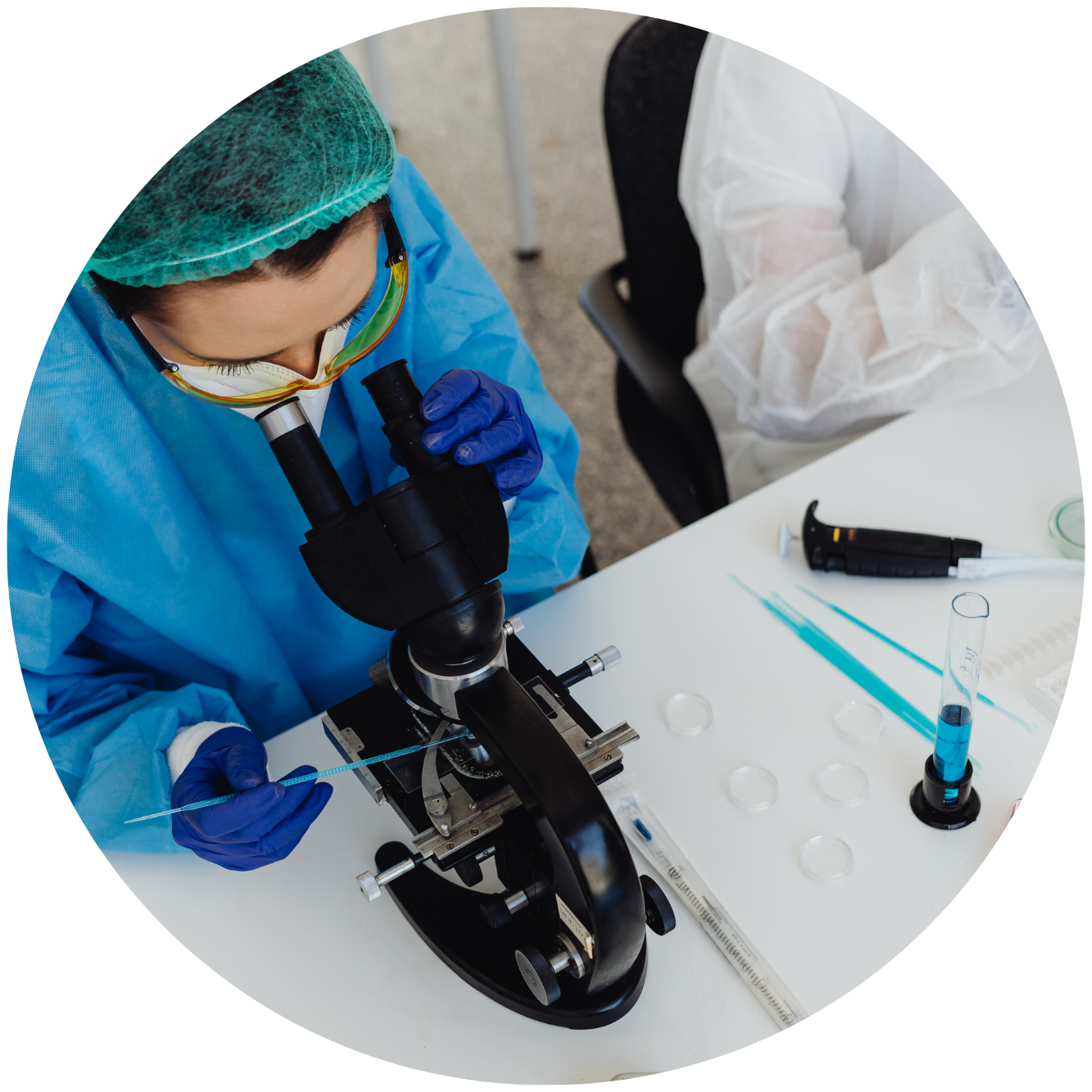 A scientist in a blue lab coat, gloves, and protective goggles looking through a microscope with a pipette in hand that demonstrates the pharmacogenetics testing services that this psychiatry provider offers.