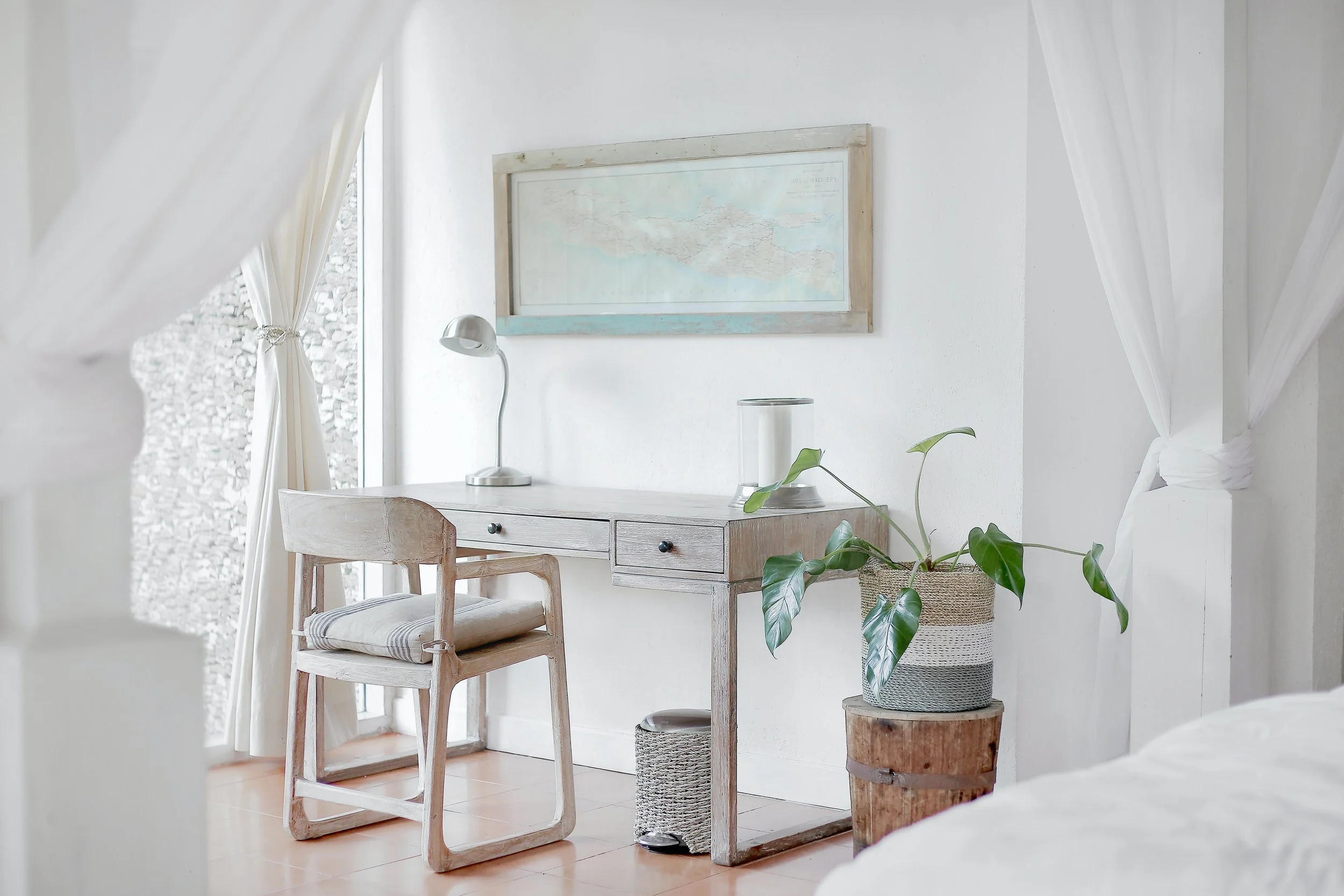A minimalist bedroom corner with white walls, a wooden desk, a wooden chair with a cushion, a small white desk lamp, a framed map on the wall, a large potted plant, and white curtains.