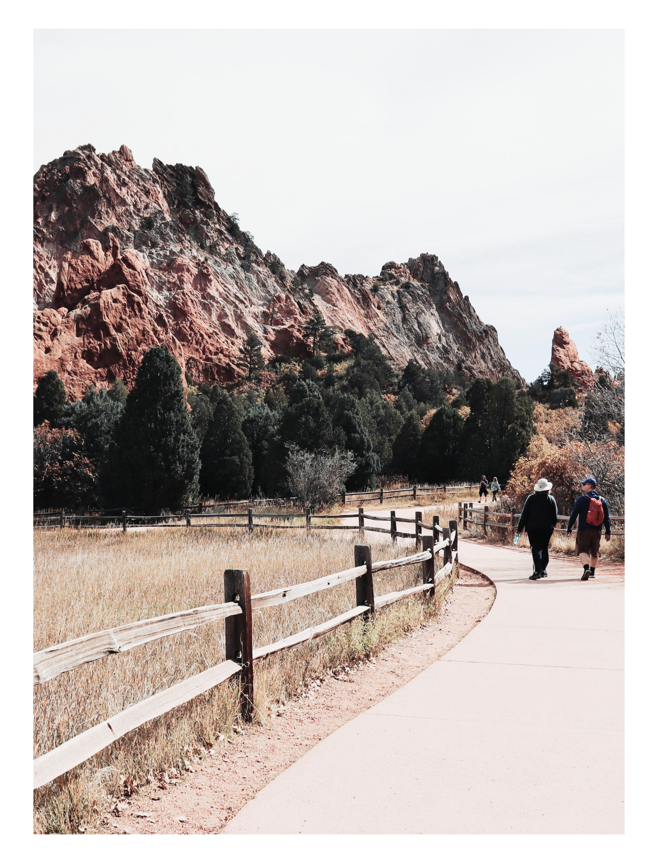 Hikers walking along a winding path with a wooden fence, surrounded by grassy fields and trees, with red rocky mountains in the background under a cloudy sky.  Representing mindfulness, resilience, mental wellness.