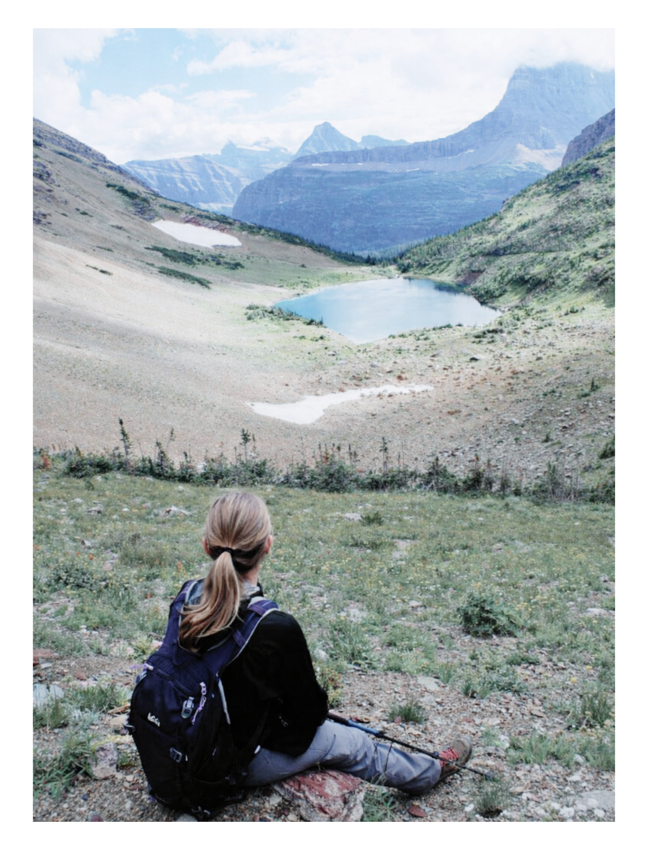 A woman with blonde hair, wearing a black jacket, gray pants, and a purple backpack, sitting on rocky ground looking at a mountain lake surrounded by mountains and snow patches.  Demonstrates resilience, doing hard things, mental wellness, Trail Mind
