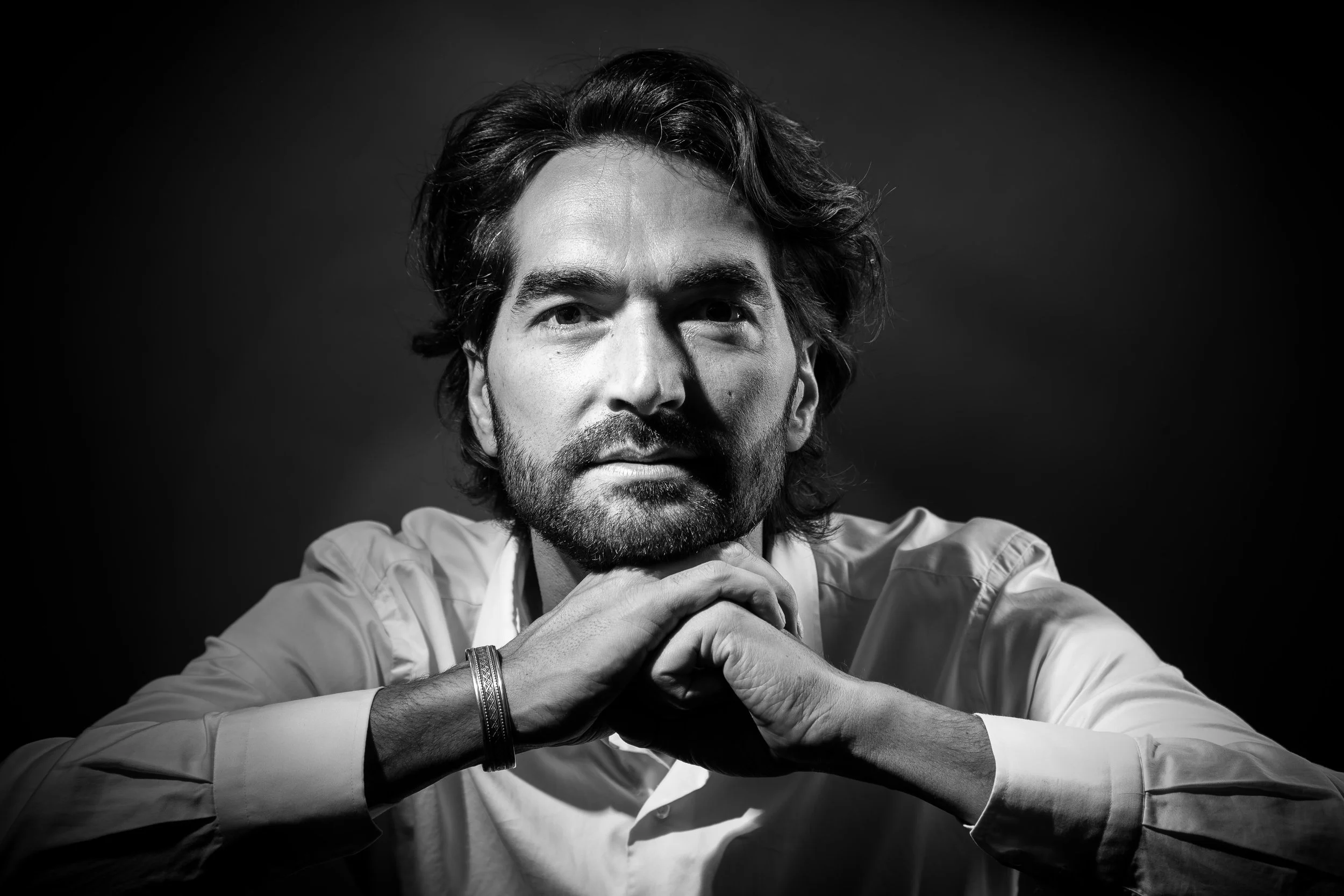Muammer Yilmaz, Black and white portrait of a man with dark hair and beard, resting his chin on his hands, wearing a long-sleeved shirt and bracelets, against a dark background.
