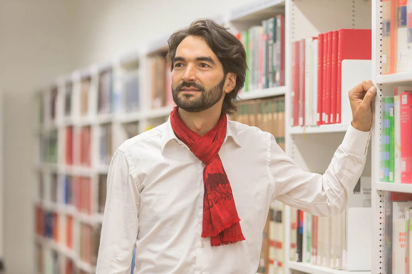 A man with dark hair and a beard, wearing a white shirt and red scarf, standing in a library aisle with shelves of books.